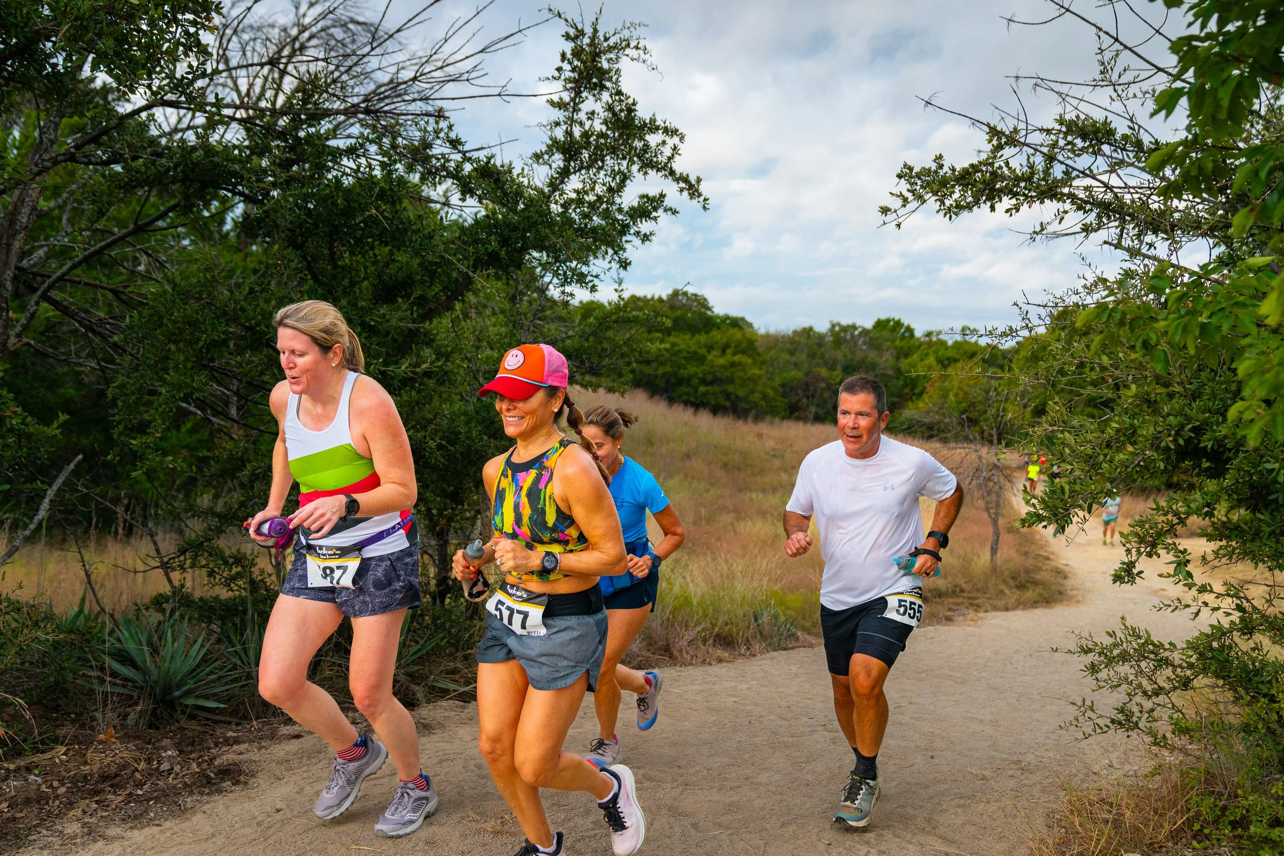 Group of four people running outdoors on a trail surrounded by trees and grass, participating in a race or marathon.
