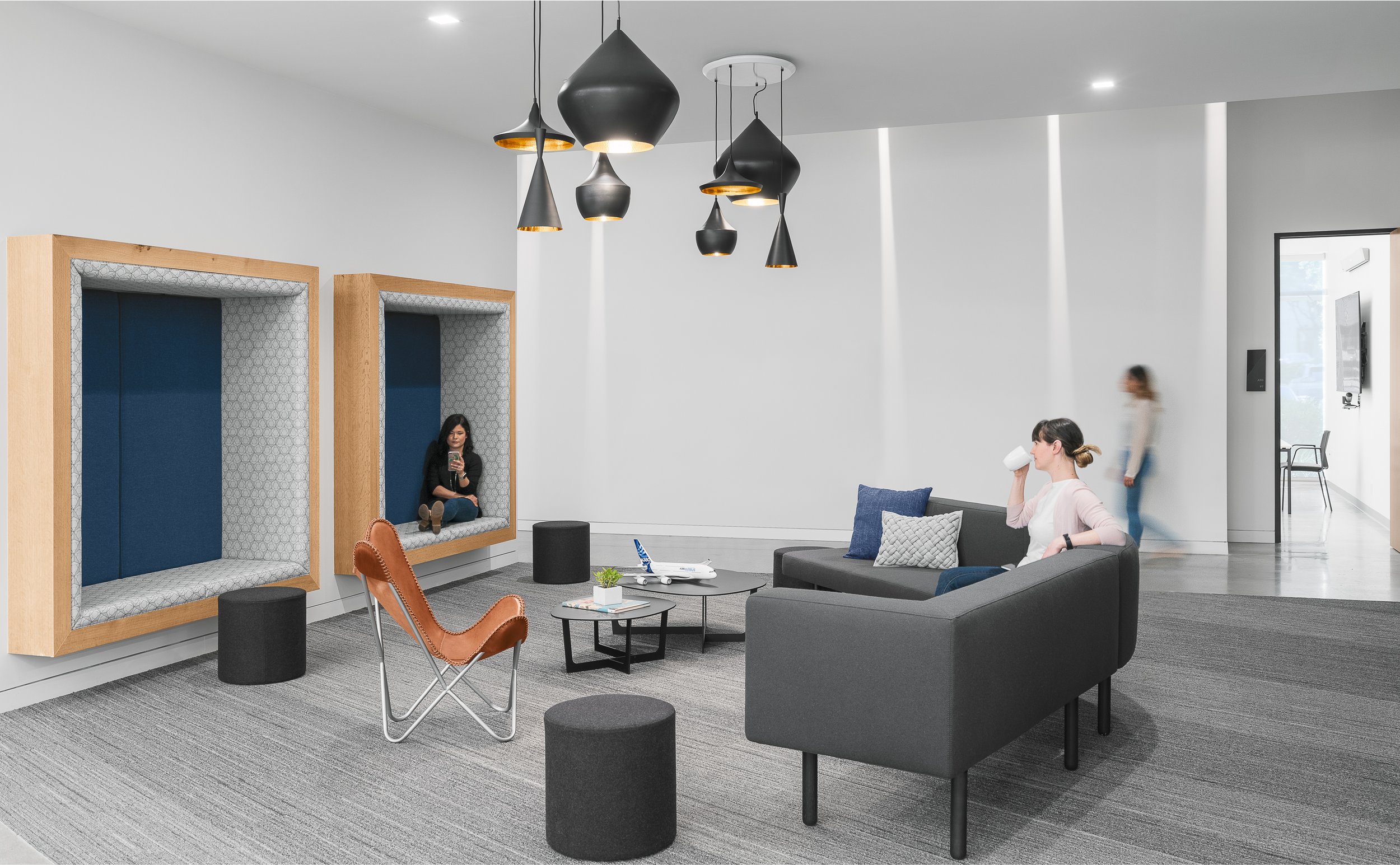 Modern office lounge with black sofa, brown chair, small black tables, and acoustic window pods, with two women sitting and drinking coffee.