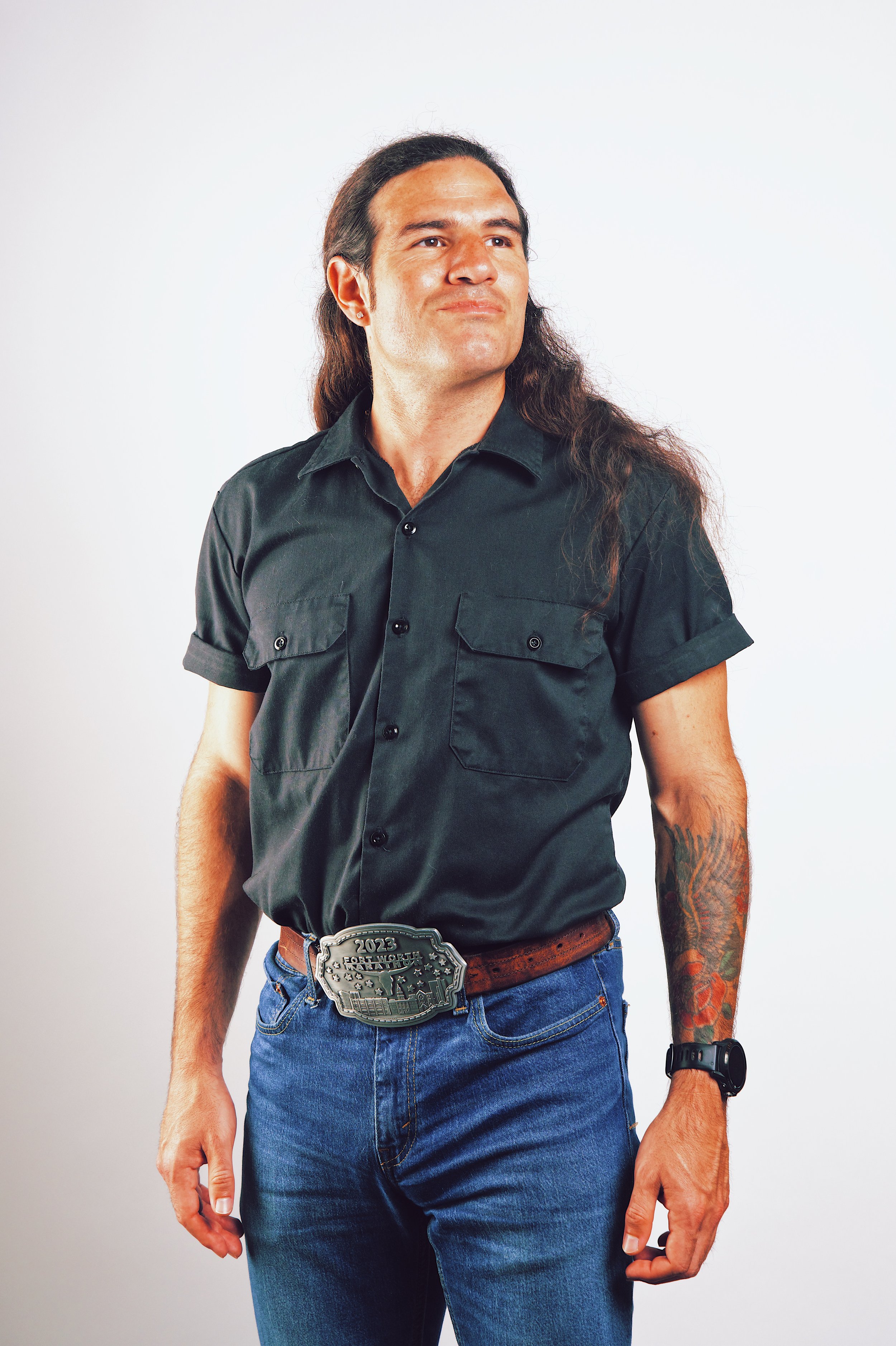 A man with long brown hair, wearing a black short-sleeve button-up shirt, blue jeans, a leather belt with a large silver belt buckle, a black wristwatch, and sporting colorful tattoos on his left arm, standing against a plain white background.