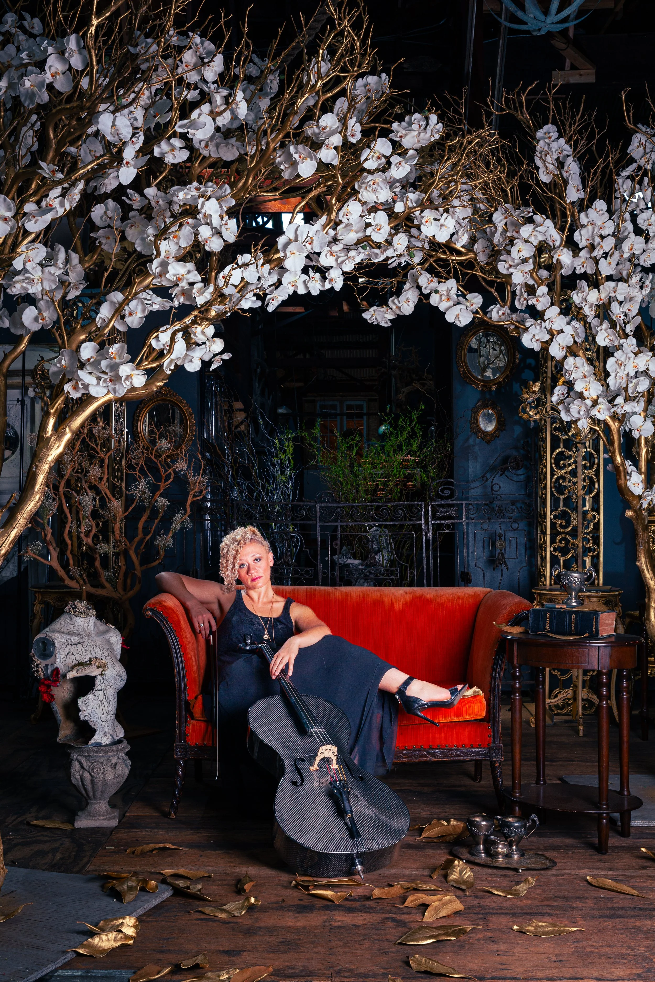A woman with curly blonde hair sitting on a red vintage sofa, holding a guitar case, in an artistic setting with large white flowers and ornate decor.