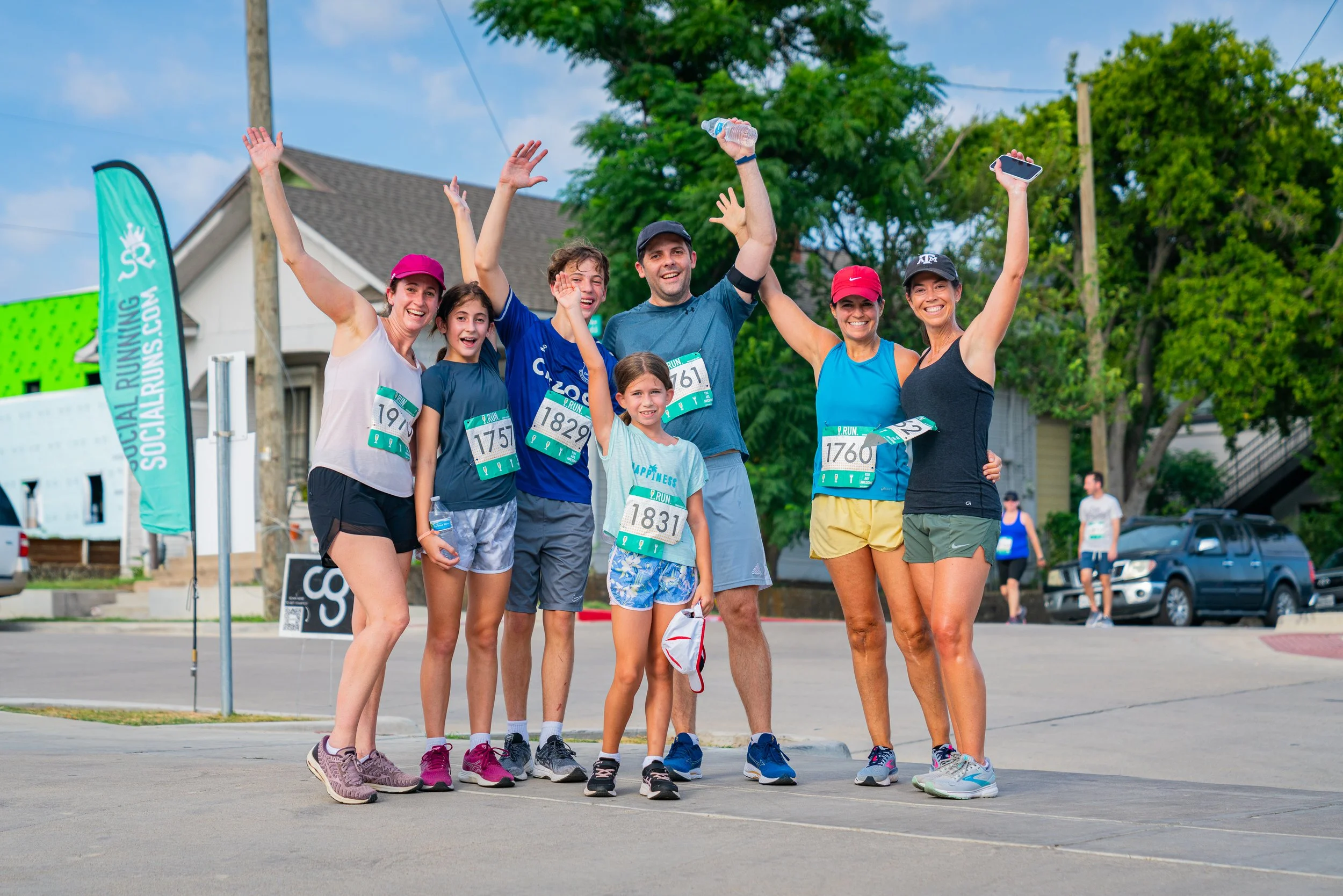 Group of seven runners celebrating after a race, standing outdoors on a city street, with green trees and houses in the background, some holding water bottles and smiling.