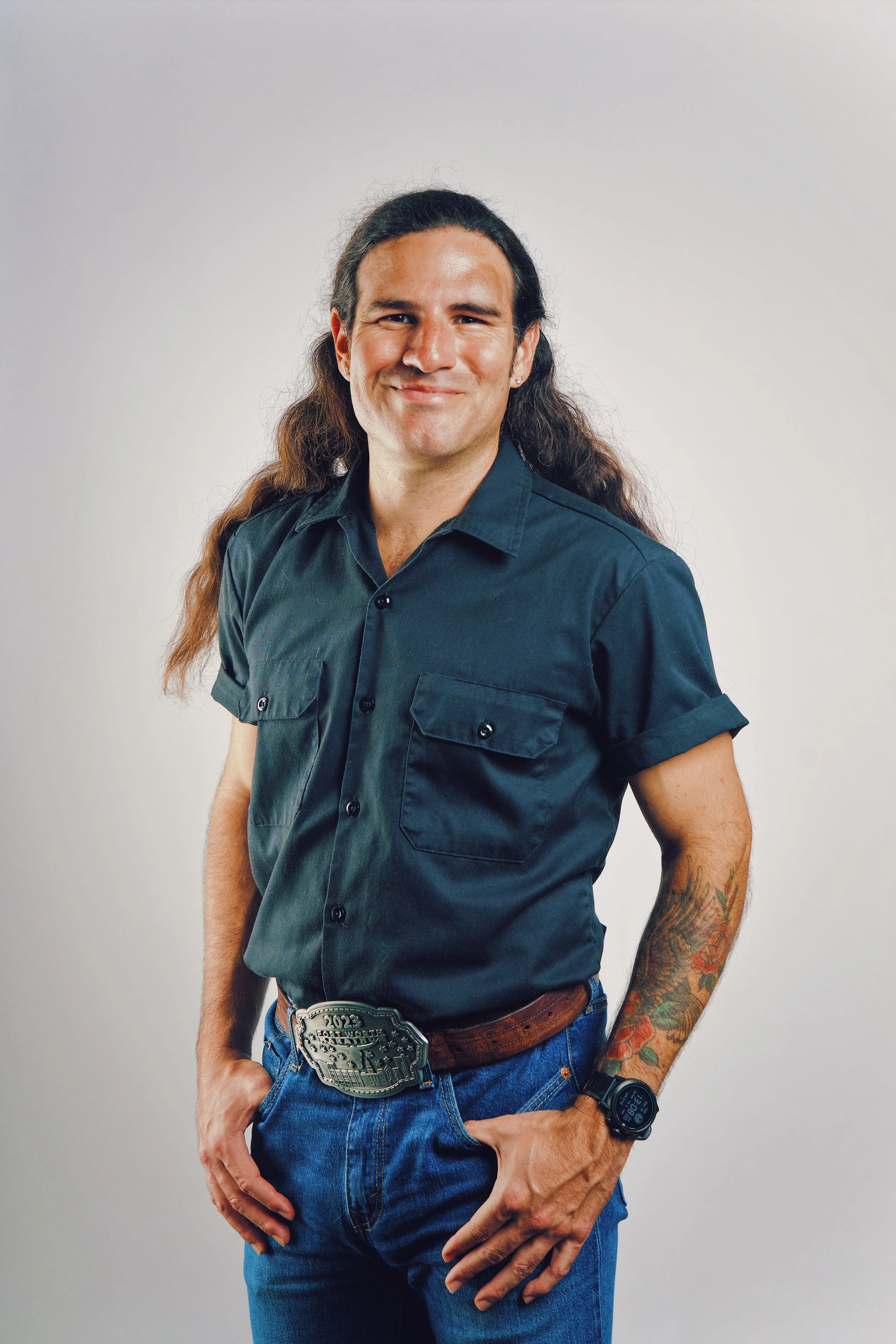 Man with long hair, wearing a black short-sleeve shirt, jeans, and a belt with a large silver buckle, standing against a plain background.
