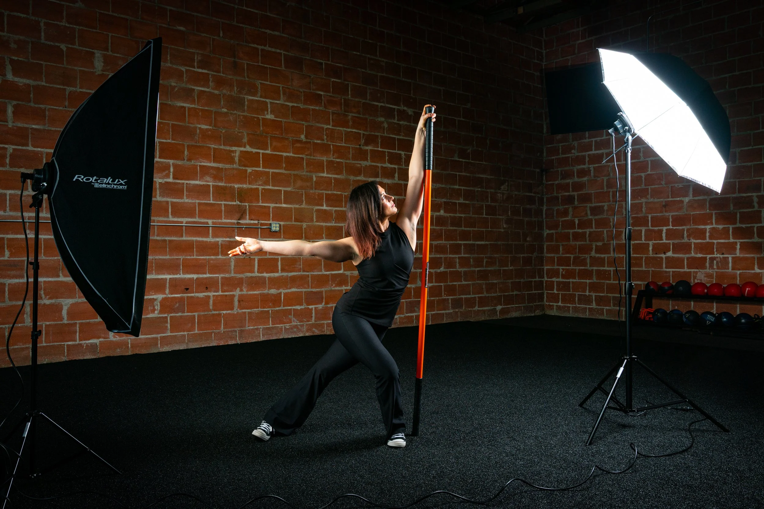 Woman practicing pole vault inside a studio with brick walls, professional lighting setup, and a gym rack with weights in the background.