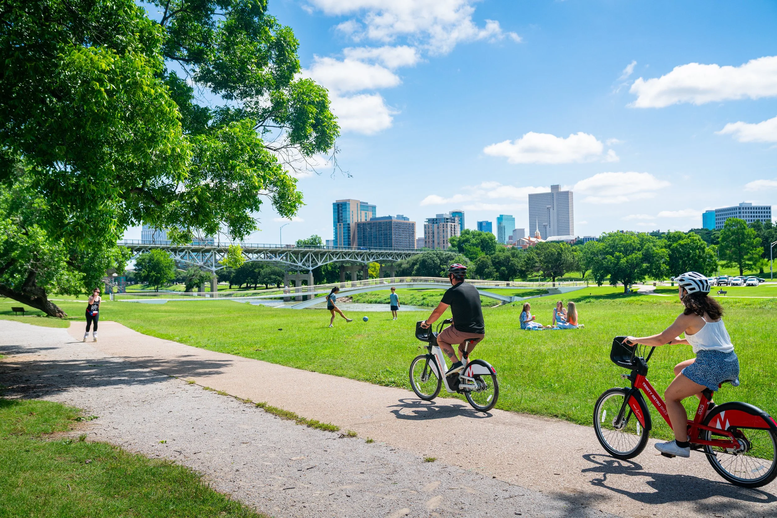 A park with children and adults enjoying outdoor activities on a sunny day. Two people ride bikes, some children play soccer, and a group of girls sit on blankets. Green trees and a city skyline with tall buildings and a bridge are visible in the background.