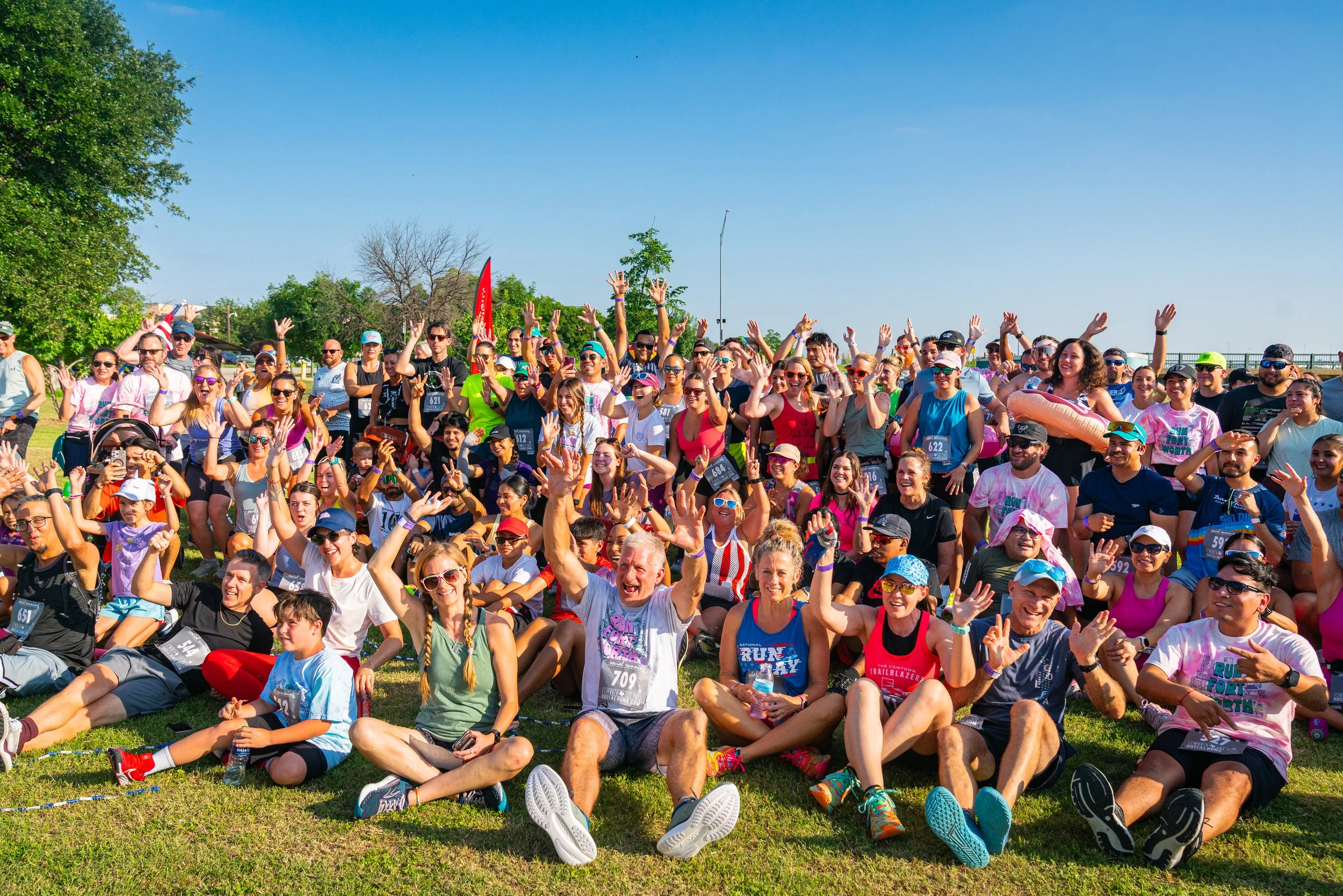 A large group of people participating in a fun run event outdoors on a sunny day, sitting and standing on grass, smiling and waving at the camera.