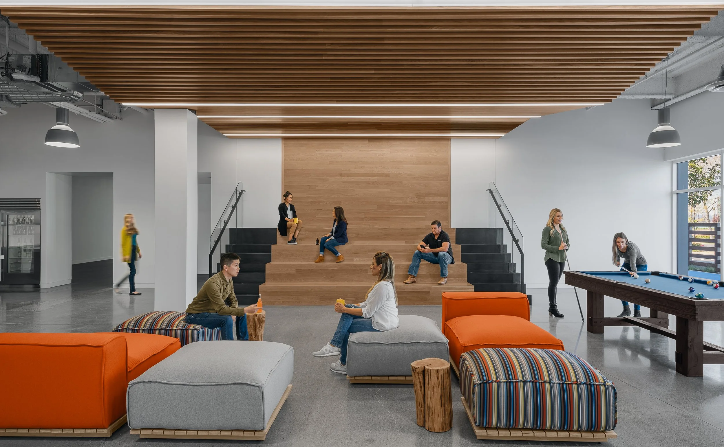 Modern lounge area with people sitting and playing pool, some on steps, some on seating, with large window and wooden ceiling.