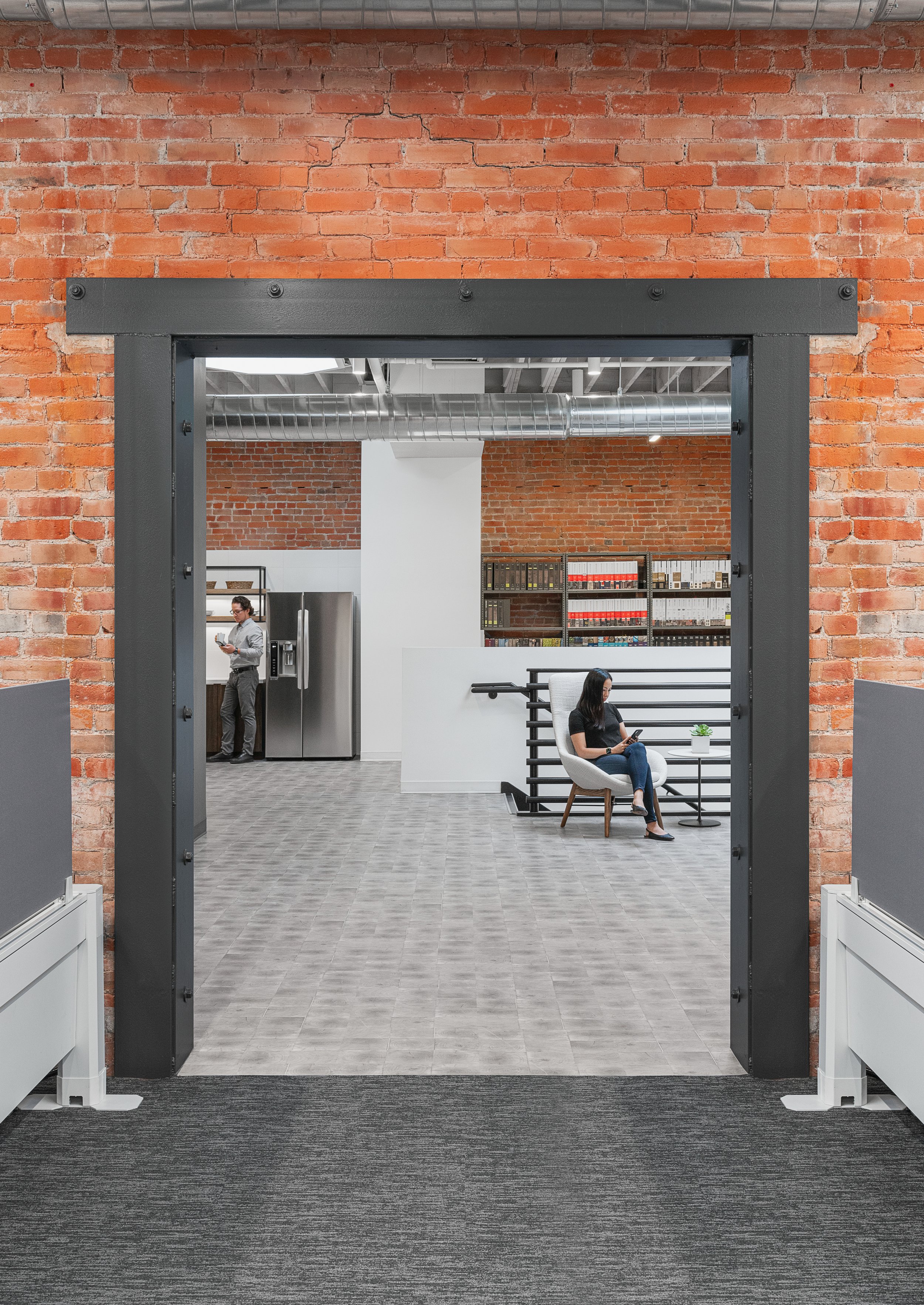 View through a doorway into an office space with exposed brick walls, a woman sitting in a chair on the right using a tablet, a man on the left near a refrigerator, and bookshelves behind the woman.