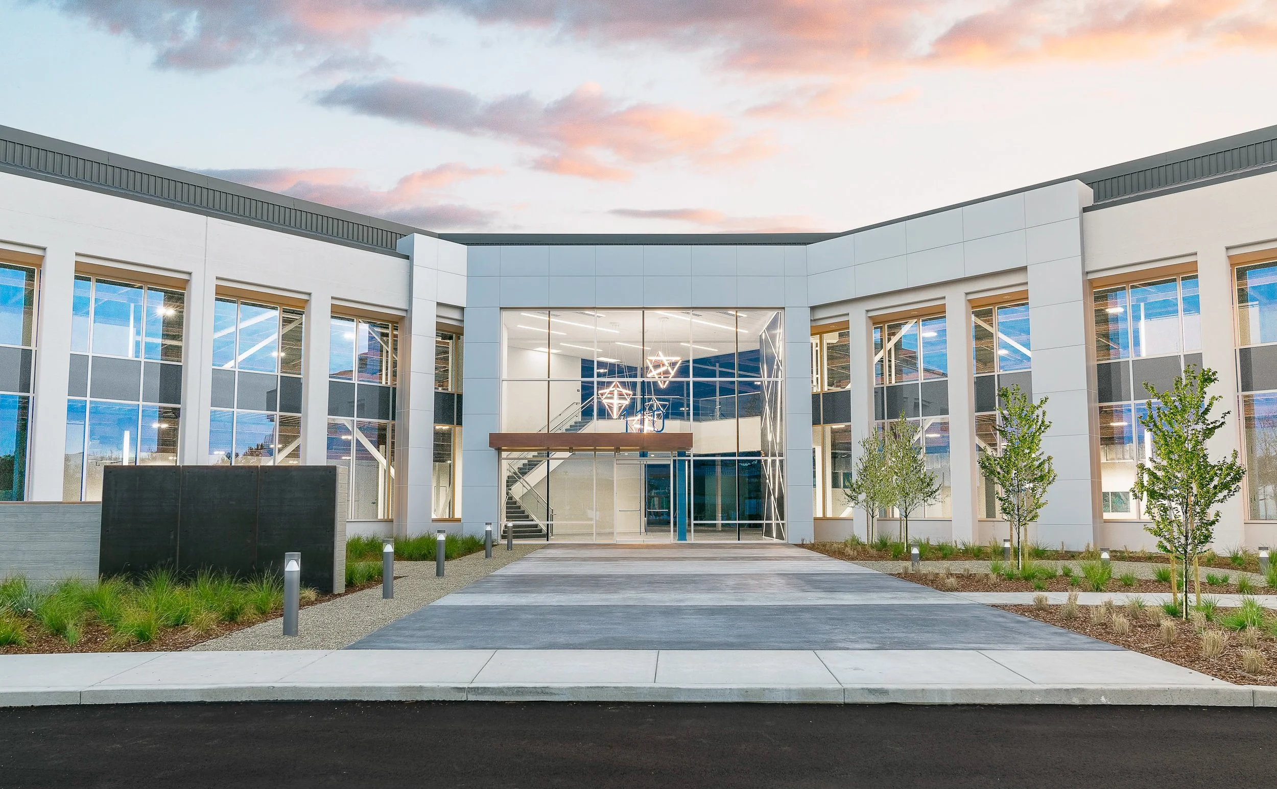 Modern commercial building with large glass windows, driveway, and landscaped surroundings at dusk.