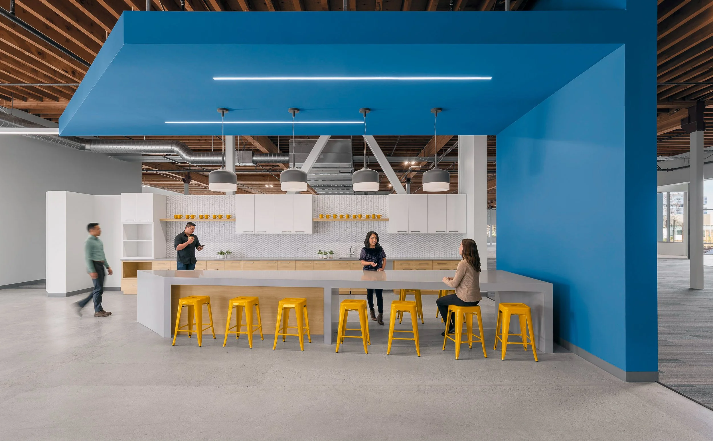 Modern kitchen area with a large white counter, yellow stools, and a blue structural element overhead. Three pendant lights hang above the counter. Two women are seated and talking, a woman is standing and talking, a man is standing and looking at his phone, another man walks by in the background. The kitchen has white cabinets and a patterned backsplash.