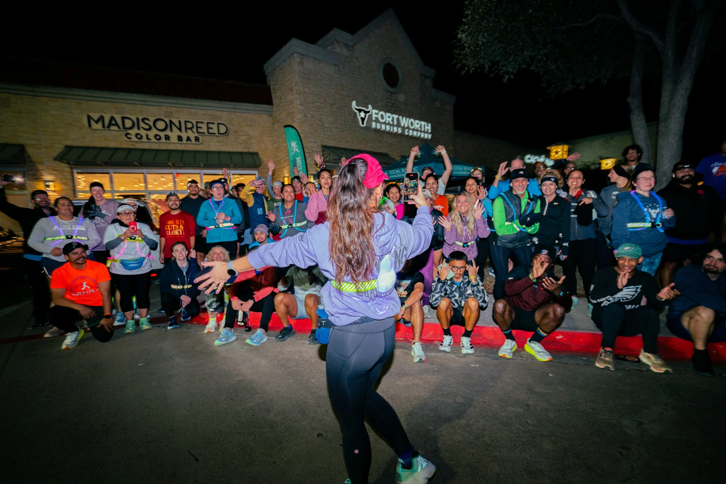 Nighttime outdoor event with a woman leading an exercise or dance session in front of a large group of diverse people, many wearing athletic clothing, in front of a building with signs for Madison Reed Color Bar and Fort Worth Brewing Company.