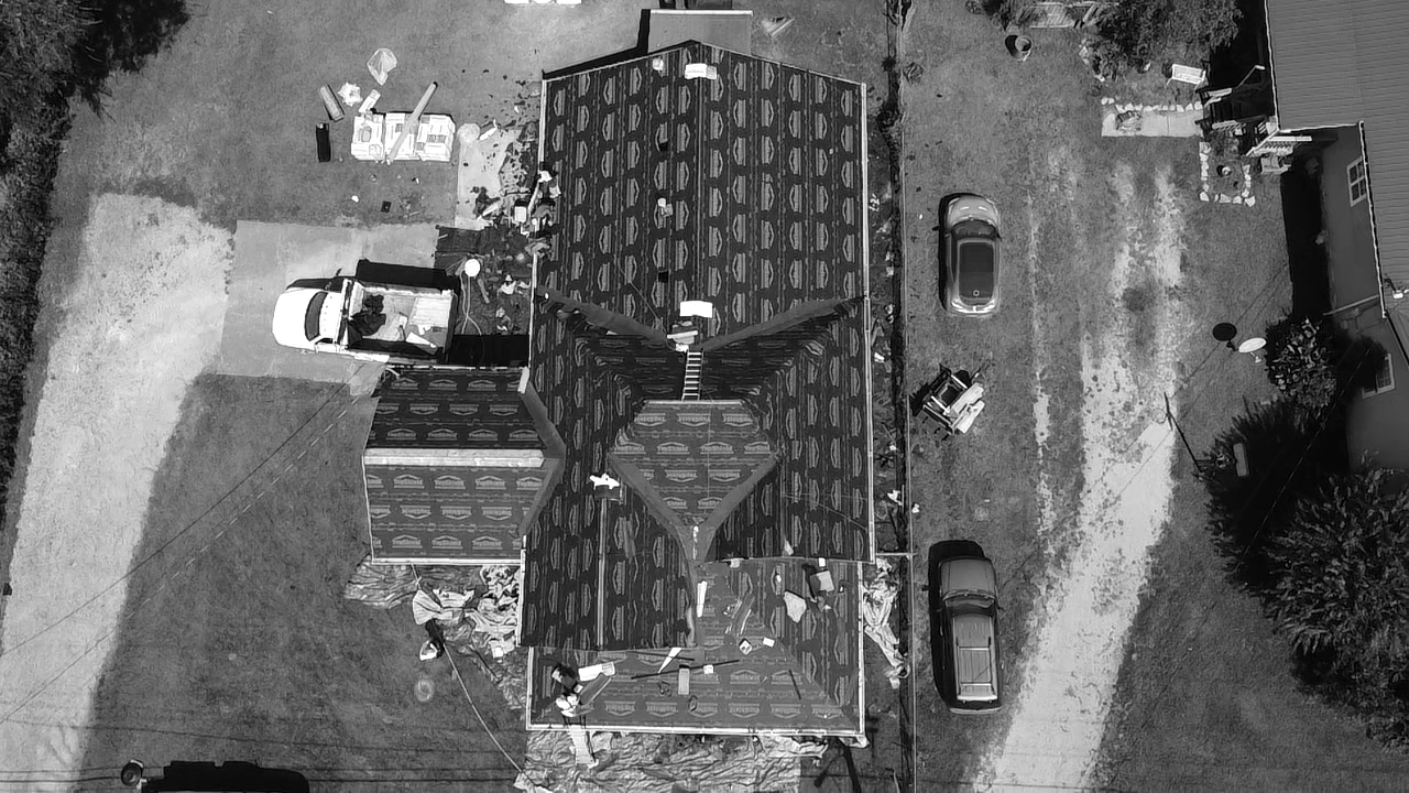 An aerial view of a house under construction with workers installing roofing, a white truck parked nearby, and two parked cars on the gravel driveway.