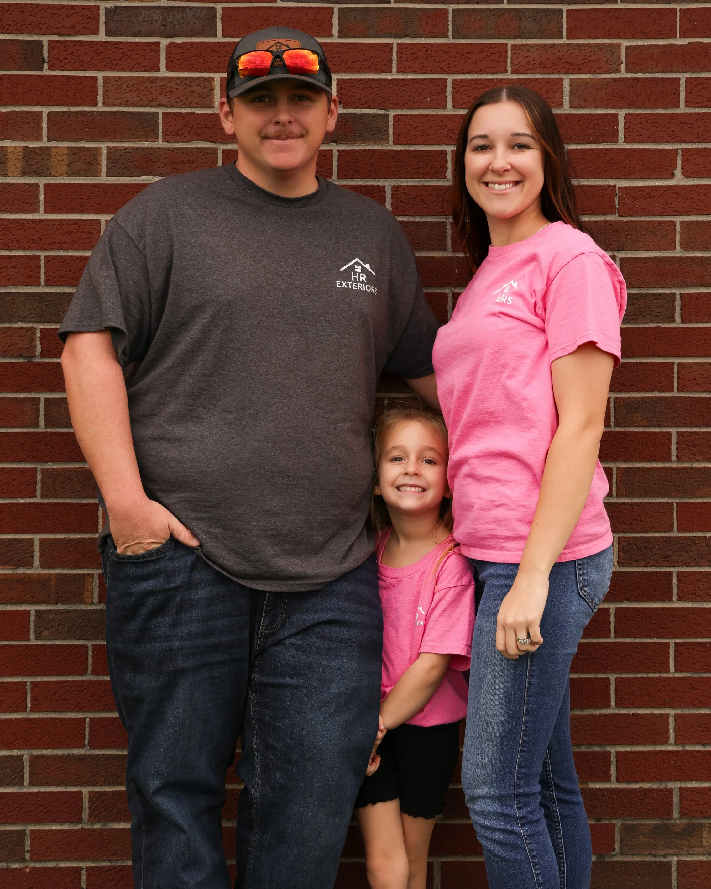 A family of three standing against a brick wall, smiling. The man on the left is wearing a dark gray t-shirt, a baseball cap, and sunglasses on his cap. The little girl is between them, wearing a pink shirt and black shorts, smiling widely. The woman on the right is wearing a pink t-shirt and blue jeans, smiling at the camera.