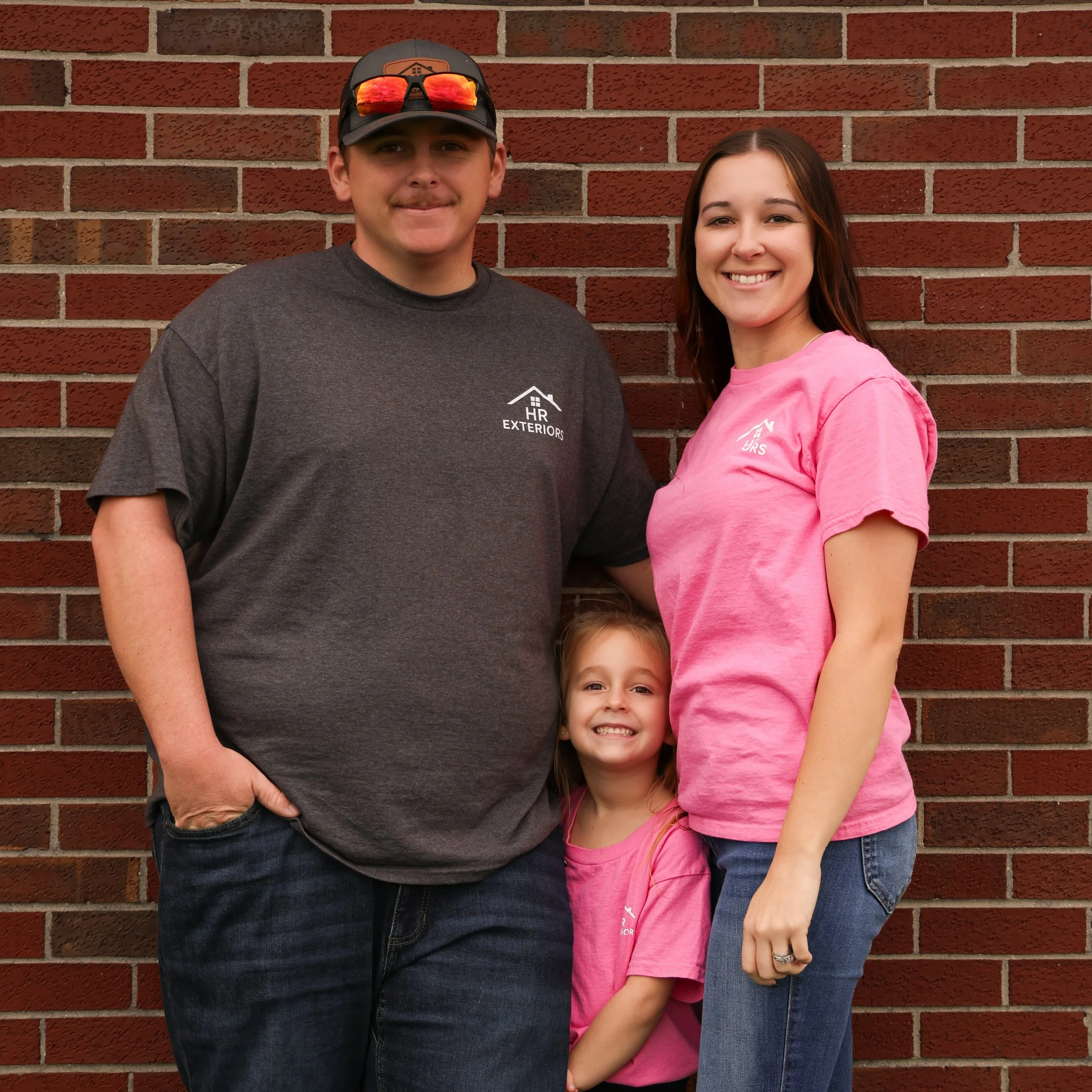 A smiling family of three standing in front of a brick wall. The man is wearing a dark T-shirt with a logo that reads 'HR Exteriors' and a baseball cap with sunglasses resting on top. The woman is wearing a pink T-shirt with a logo and blue jeans. The young girl, standing between them, is also wearing a pink T-shirt and is smiling.