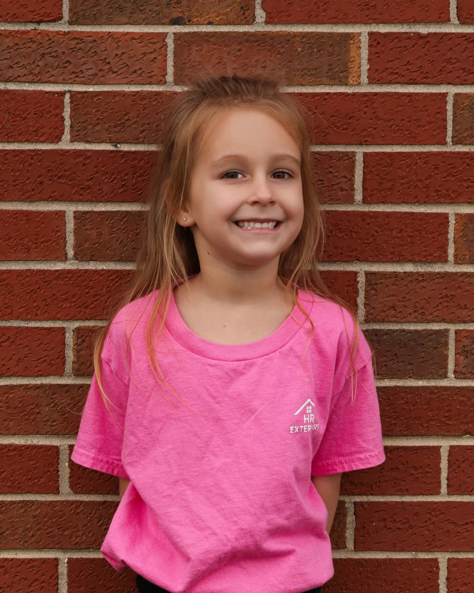 A young girl with long red hair smiling and standing in front of a red brick wall. She is wearing a pink t-shirt with white text and a small house logo.