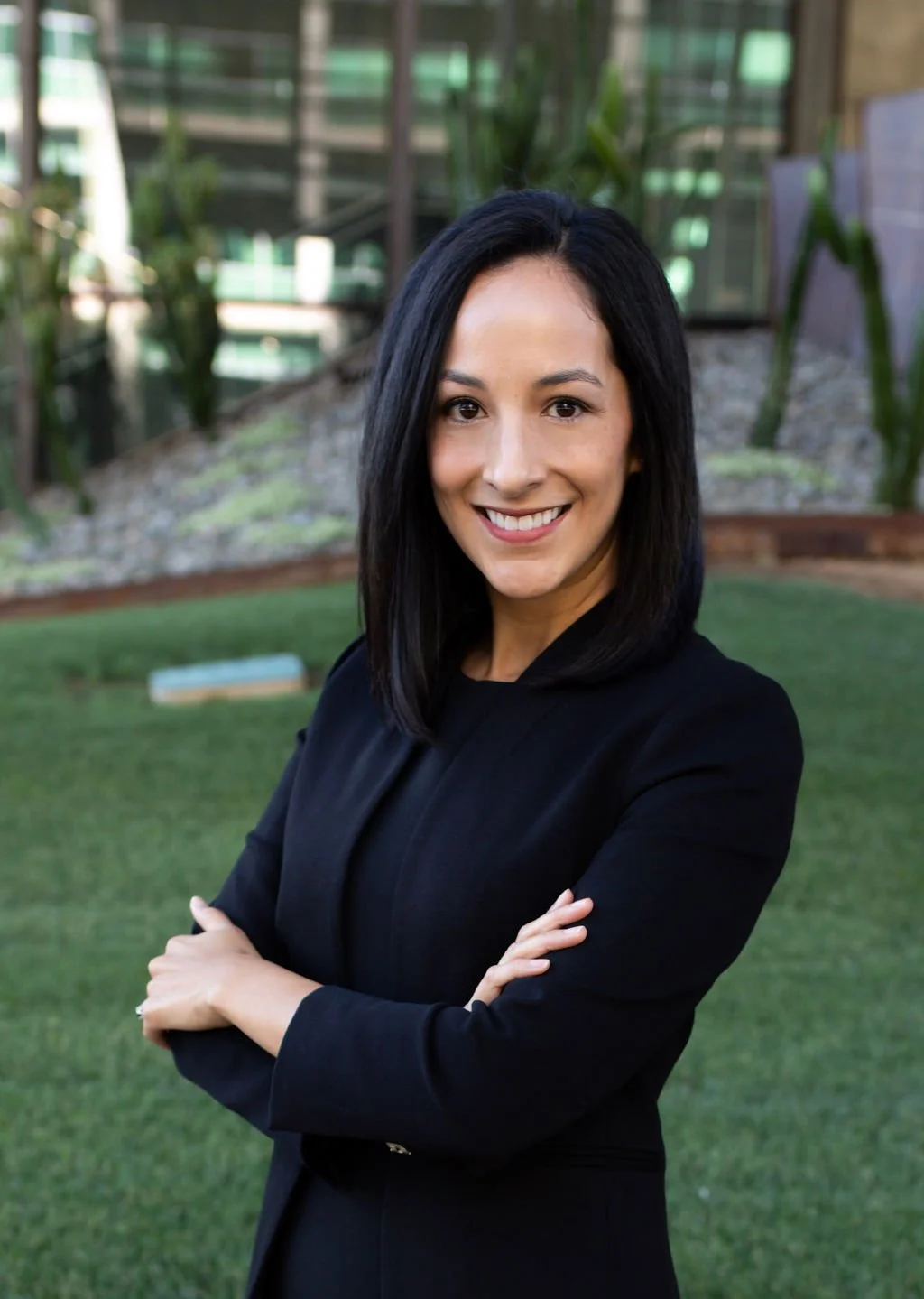 A woman with black hair smiling outdoors, wearing a black blazer, with a grassy area and modern building in the background.