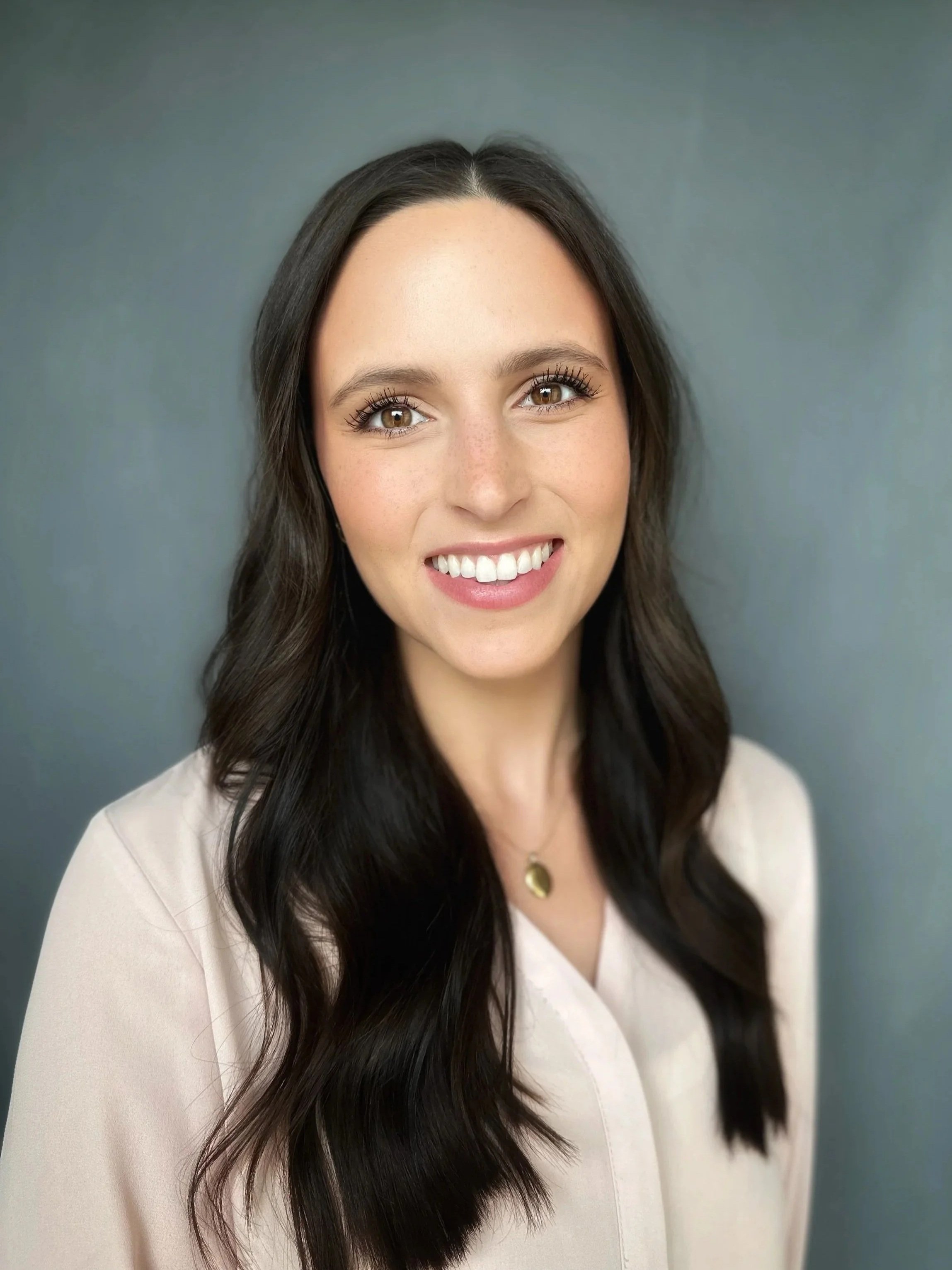 Close-up portrait of a young woman with long, dark, wavy hair, smiling, wearing a light-colored top and a gold necklace, against a plain, muted gray background.