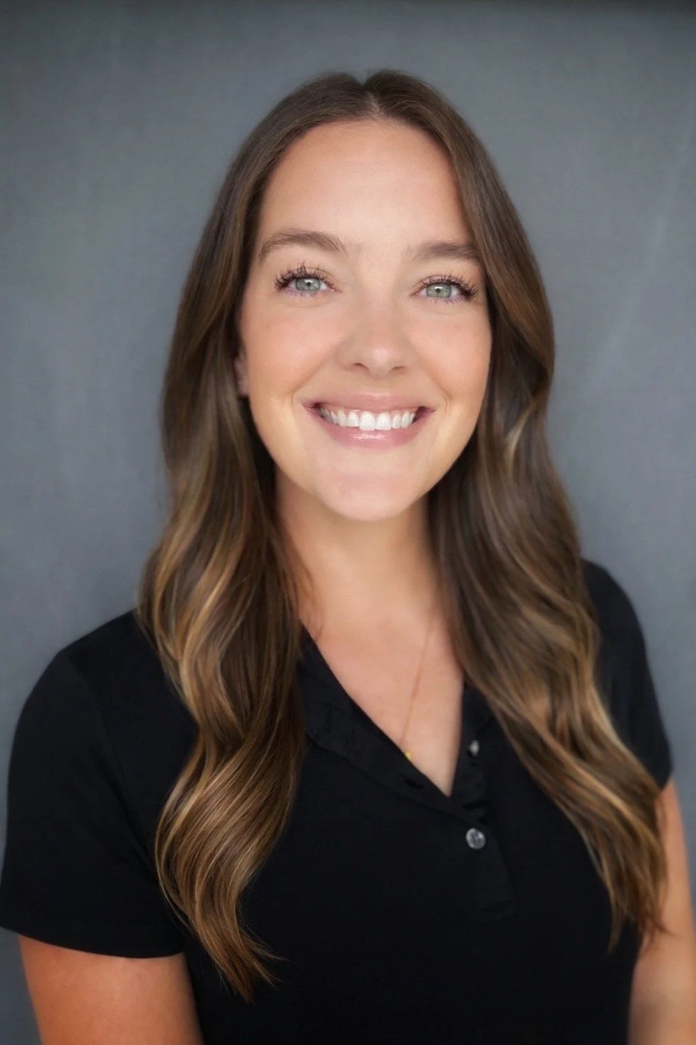 Portrait of a woman with long wavy brown hair, wearing a black shirt, smiling, against a gray background.