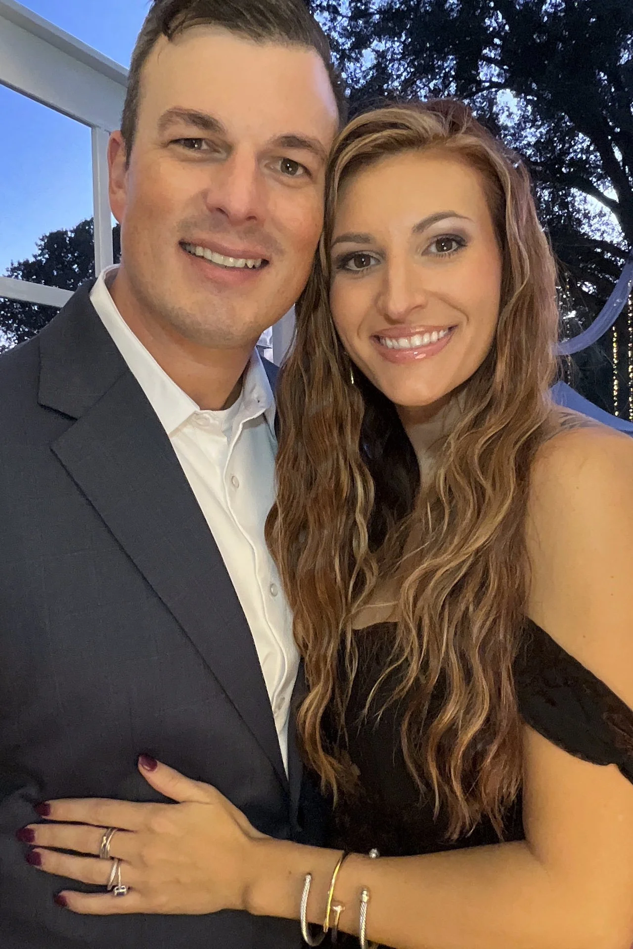 A smiling man and woman posing together outdoors during the evening. The man is wearing a dark suit and white shirt, and the woman has long curly hair, is wearing a black dress, and has jewelry on her wrist and fingers.