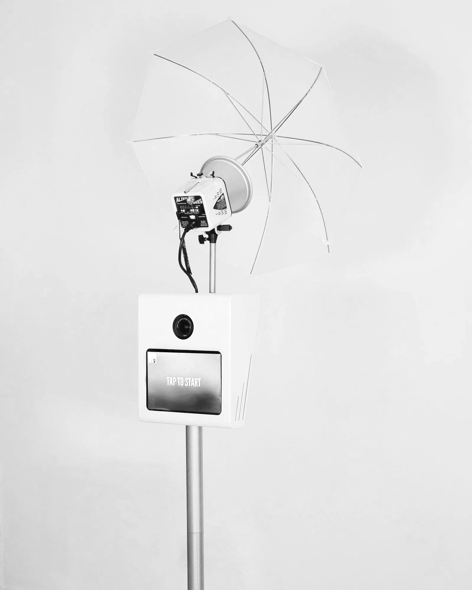 A photo of a sleek, modern photo booth setup with a large umbrella light on a stand, a professional camera, and a touchscreen display that reads 'TAP TO START'.
