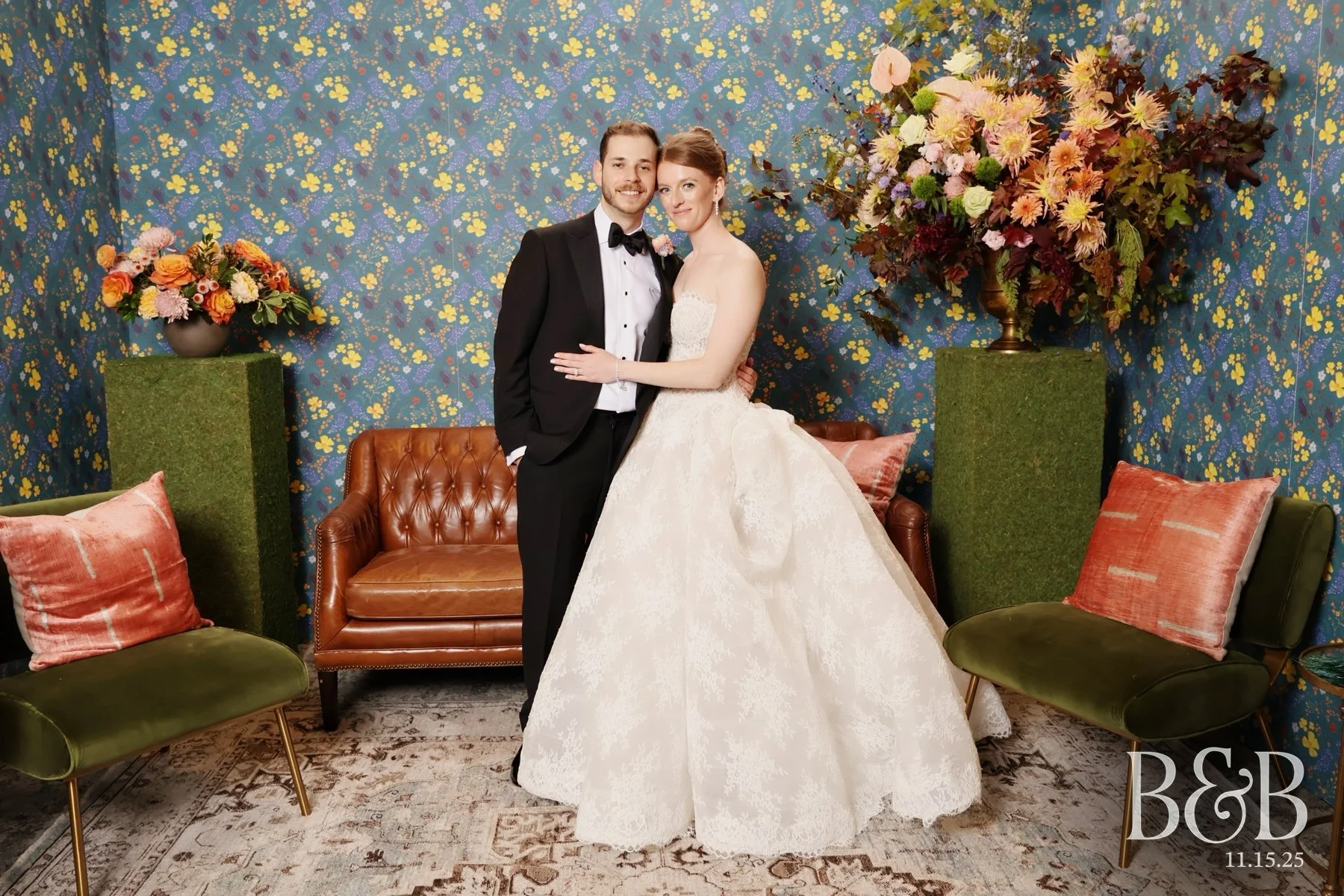 A bride and groom in wedding attire posing together in front of a floral background with colorful flowers and vintage style furniture, including a green and a brown chair and a floral arrangement on tall stands. The date 11.15.25 and initials B&B are in the bottom right corner.