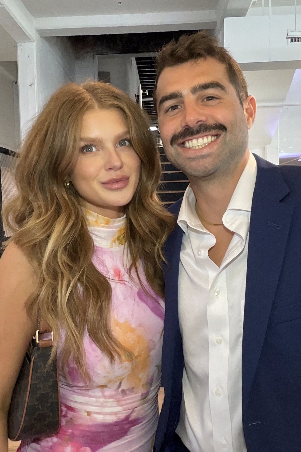 A smiling woman with long, wavy red hair wearing a colorful tie-dye dress standing next to a smiling man with short brown hair and a mustache wearing a dark blue blazer and white shirt, taking a selfie together indoors.
