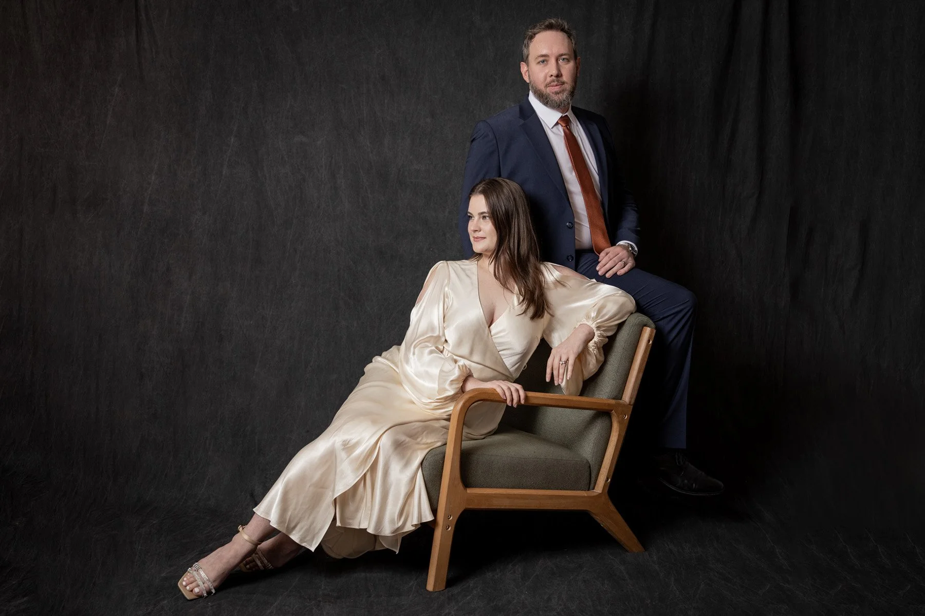 A man in a dark suit and a woman in a cream-colored satin dress pose against a dark background. The woman is seated on a wooden armchair, leaning back with one arm on the chair's armrest and the other hand resting on her lap. The man stands behind her with one knee on the chair and his hand on his thigh.