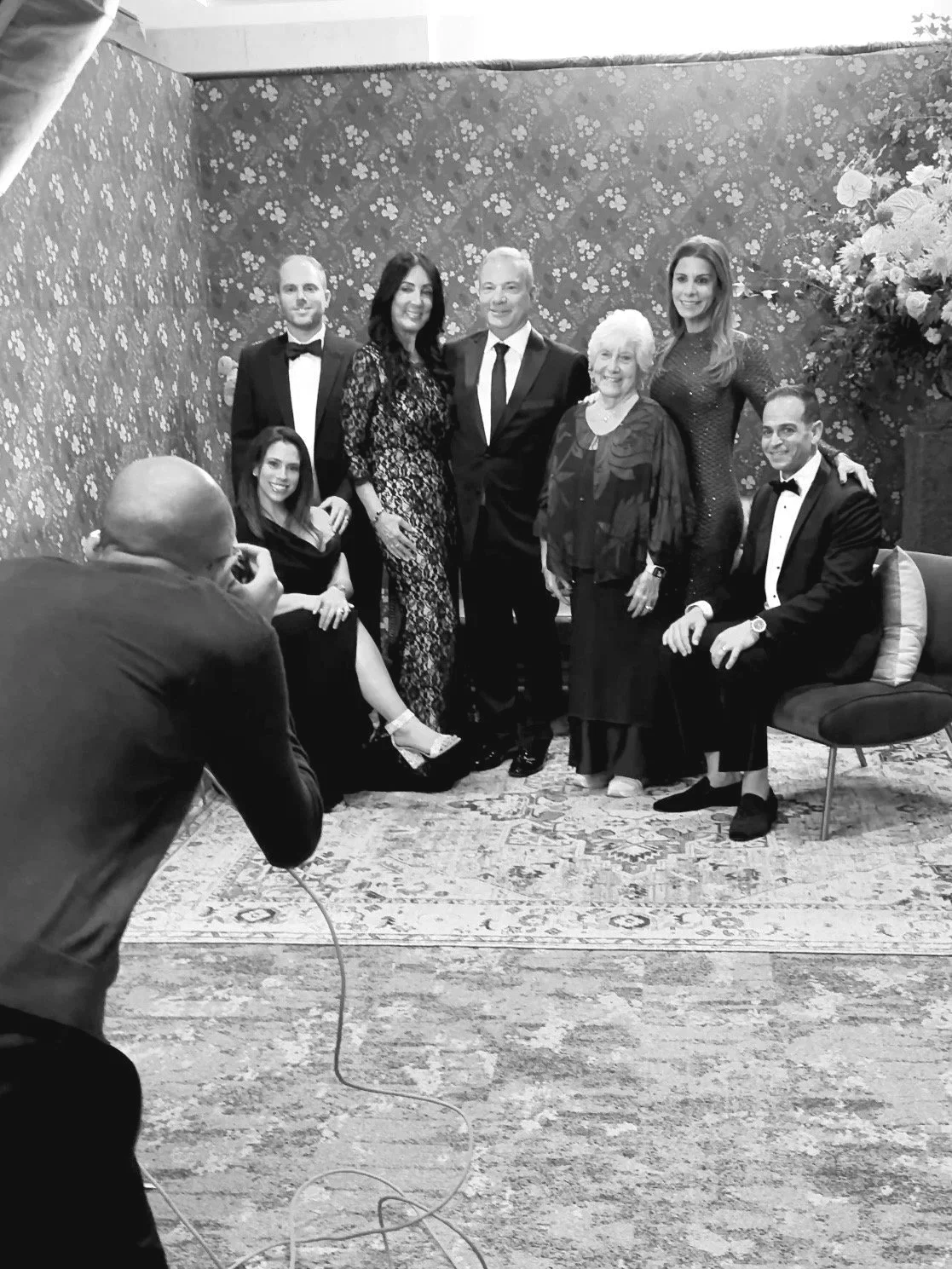 A group of eight people dressed in formal attire posing for a photo in front of a floral wallpaper background, with a photographer taking their picture from the left.