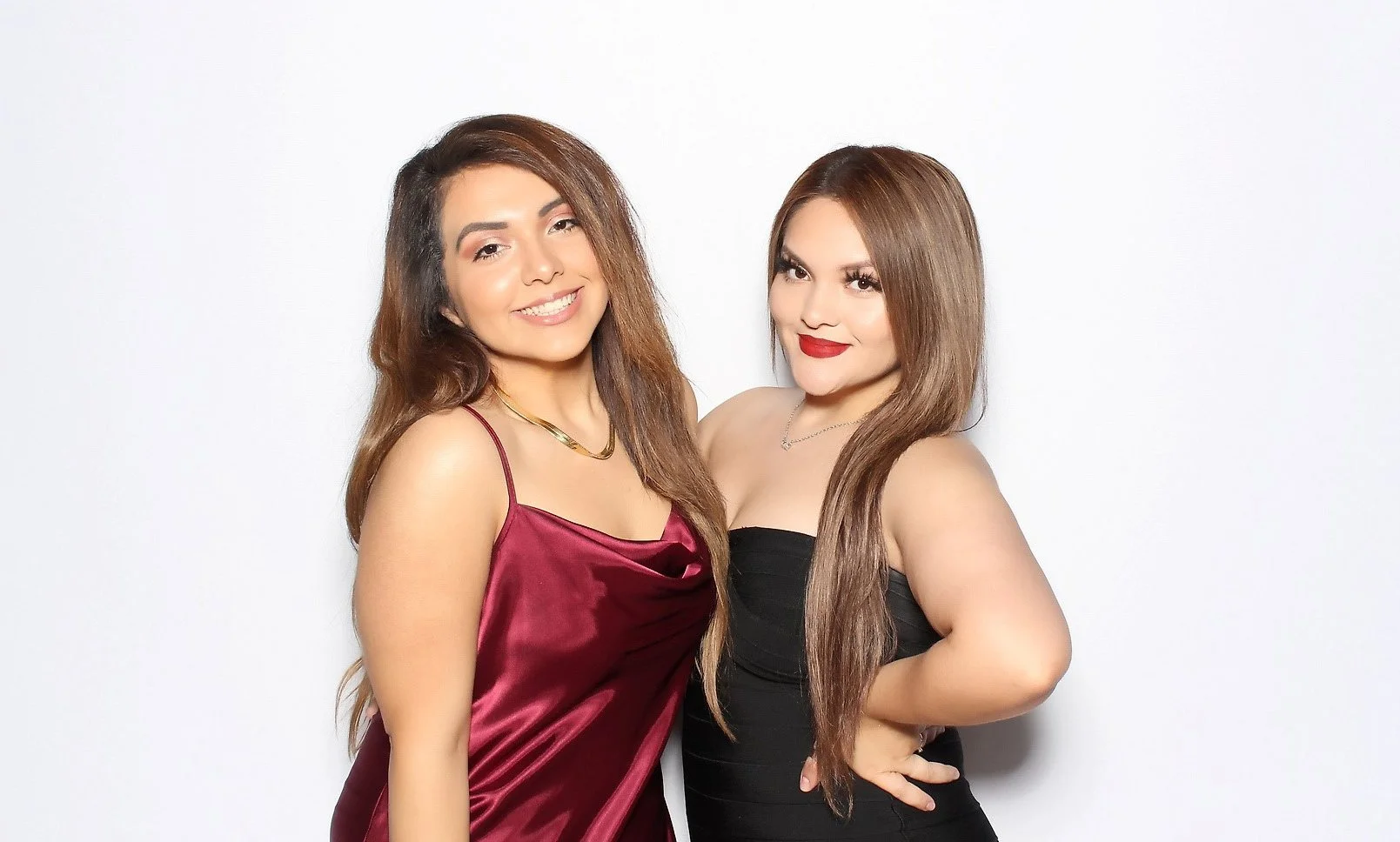 Two women in elegant dresses standing close, smiling, with white background.