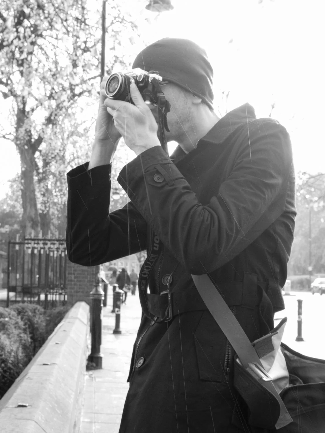 A photographer standing outdoors on a sidewalk, taking a picture with a camera, wearing a dark jacket and a beanie, with greenery and other people in the background.