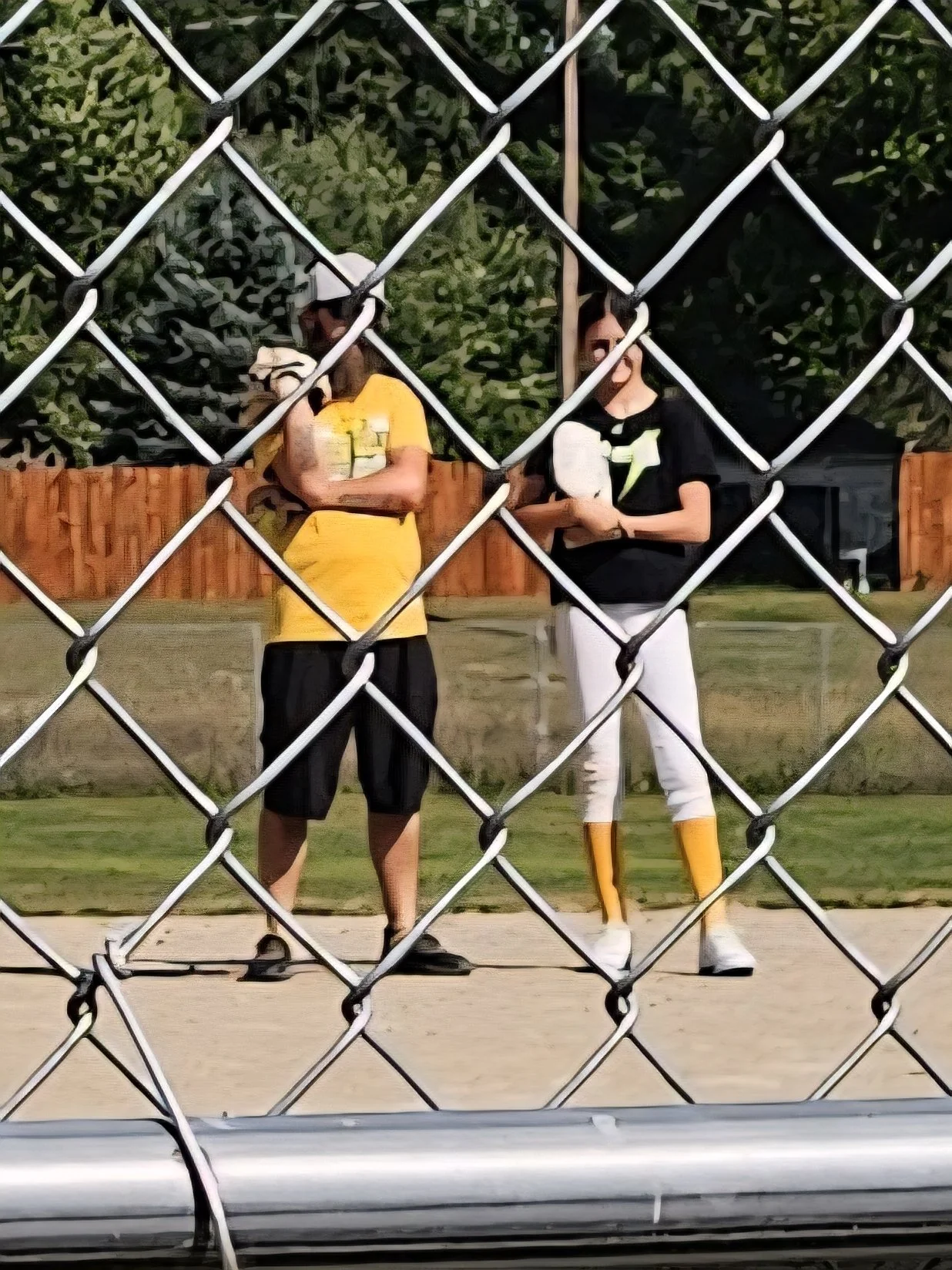 Two people standing on a sports field, viewed through a chain-link fence. One person is wearing a yellow shirt and black shorts, holding a water bottle. The other person is dressed in black and white with yellow accents, including tall yellow and white boots.