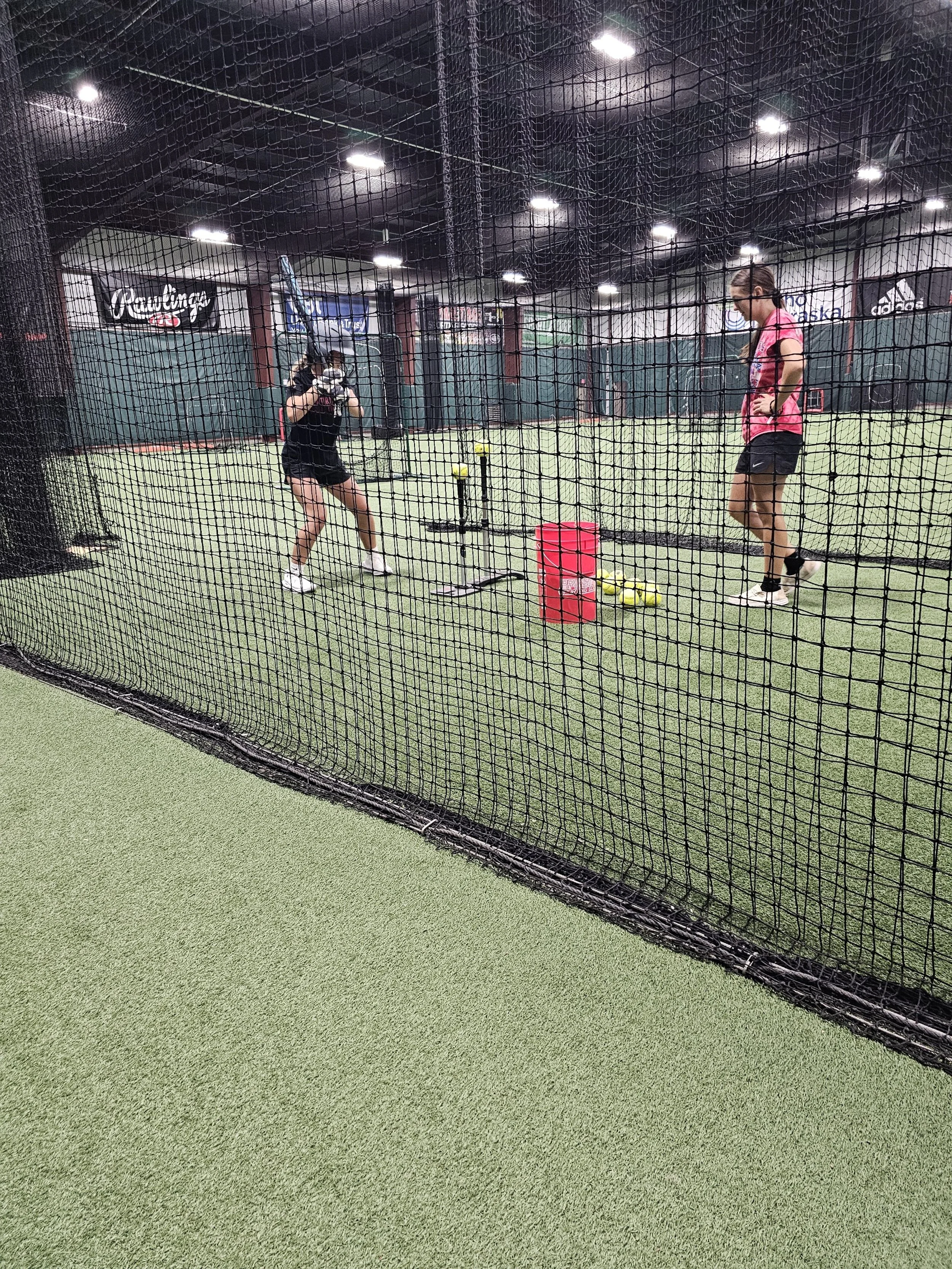 Two women practicing baseball inside a batting cage, one holding a bat and the other coaching, with tennis balls and a red crate on the ground.