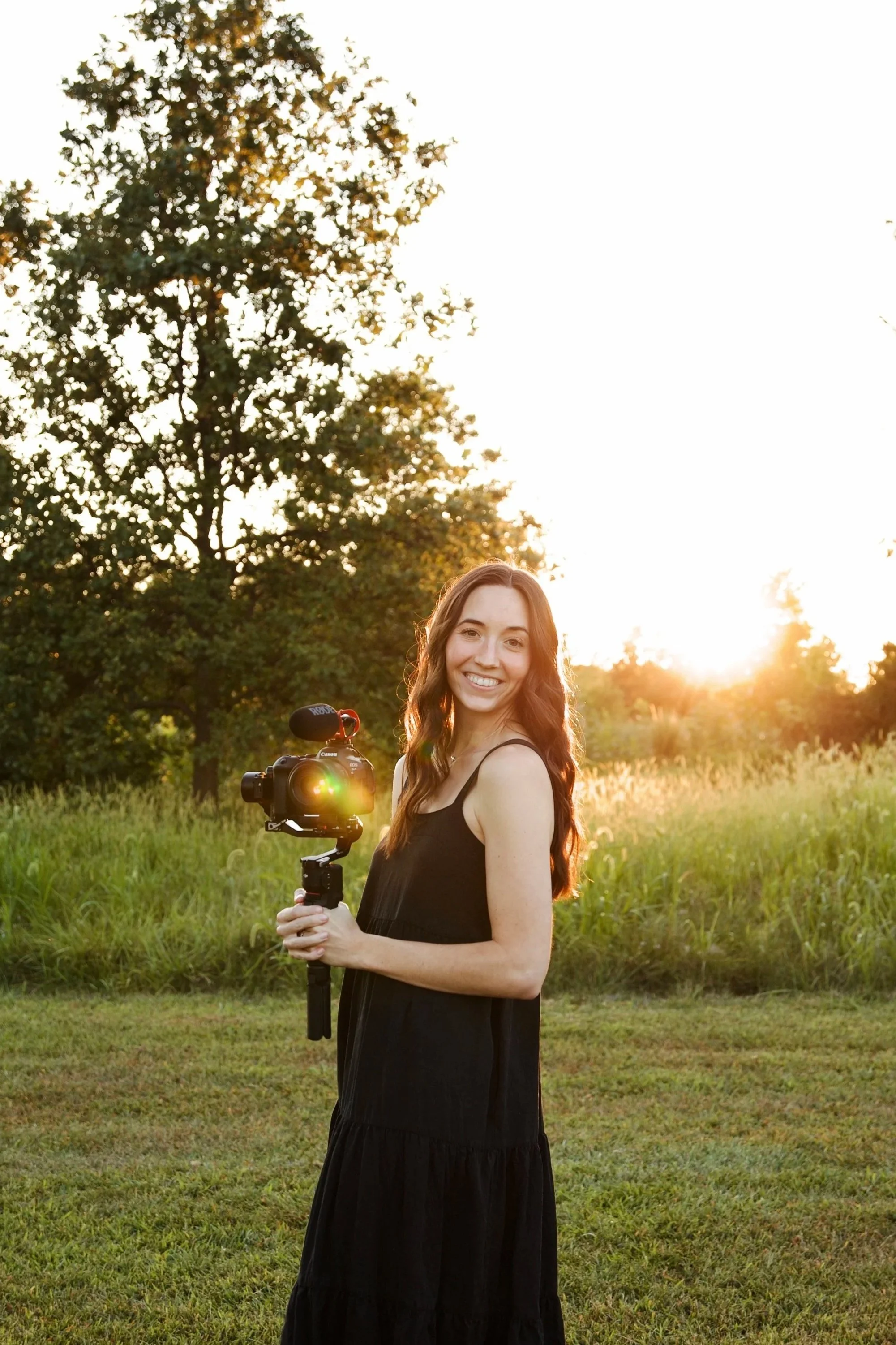 A smiling woman with long brown hair standing outdoors during sunset, holding a camera on a stabilizer, with a grassy field and trees in the background.