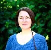 A woman with short brown hair, wearing a blue top, standing outdoors in front of green foliage, smiling at the camera.