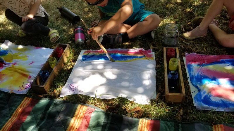 Children painting tie-dye shirts outdoors on the grass, with containers of colorful dye nearby.