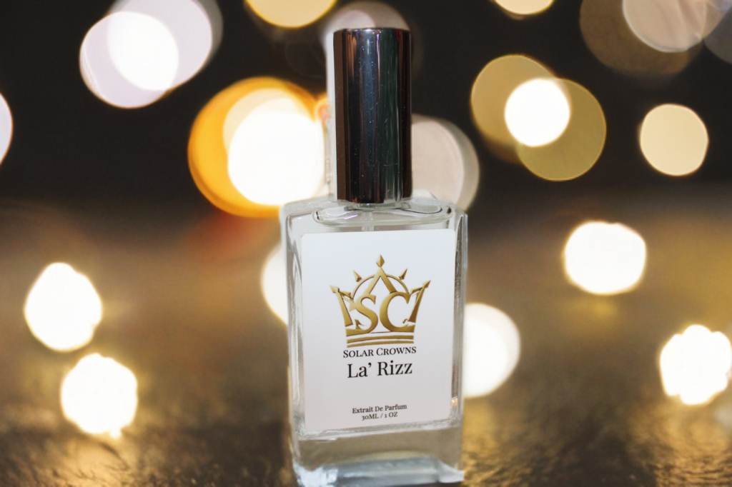 A clear glass perfume bottle with a silver cap, labeled 'Solar Crowns La Rizz', placed on a surface with blurred bright lights in the background.