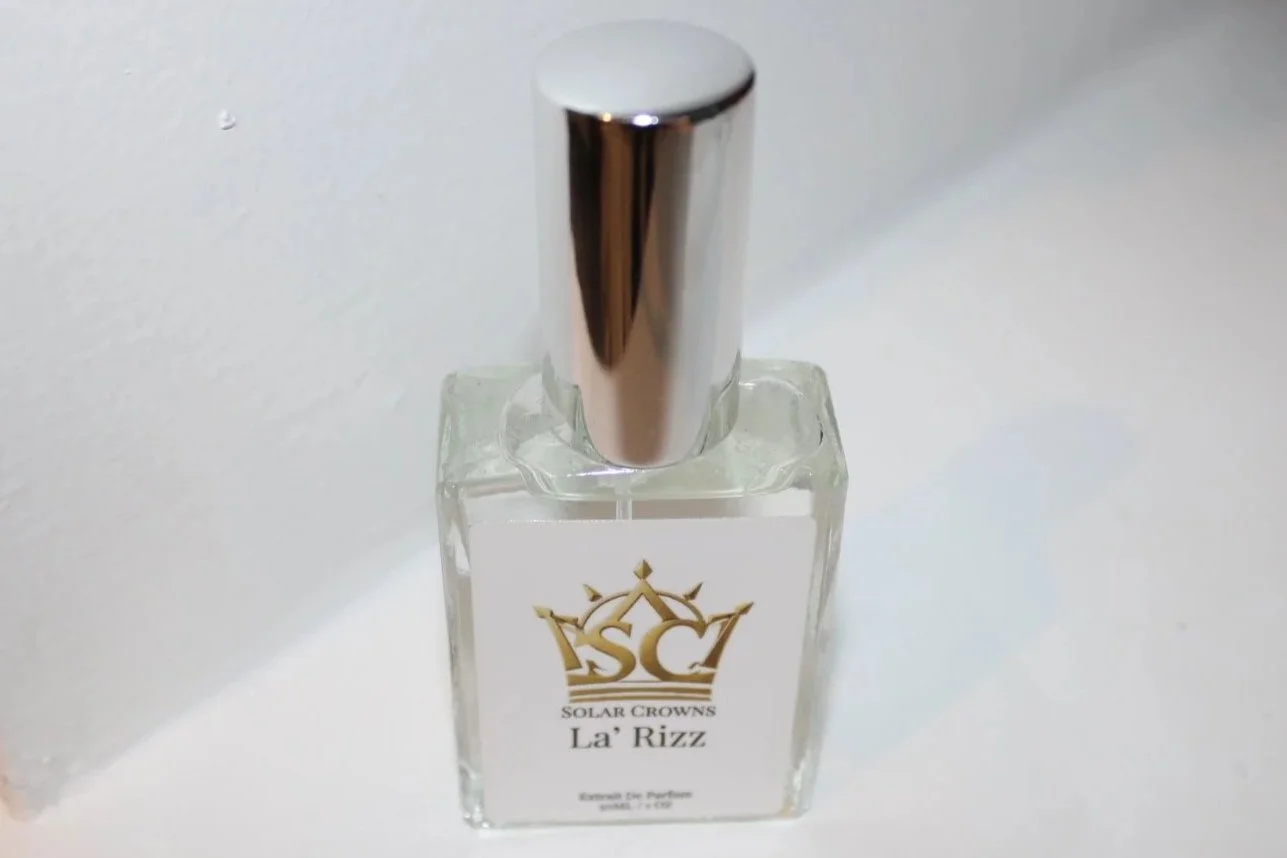 A clear glass perfume bottle with a metallic cap and a white label featuring a gold crown logo and the text 'Solar Crowns La Rizz'.