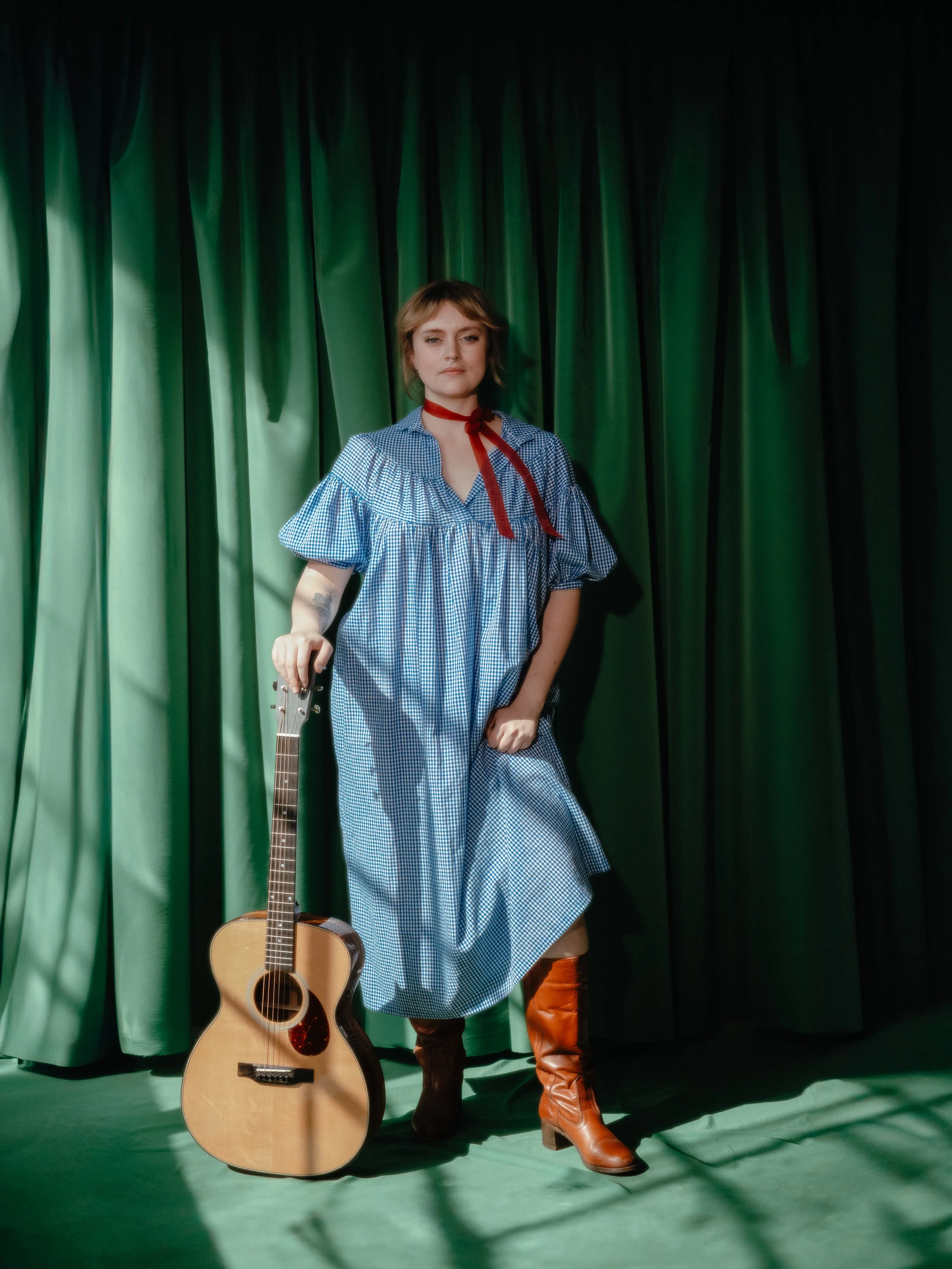 A woman standing on a stage with green curtains, holding an acoustic guitar and wearing tall brown boots, a blue and white checkered dress, and a red ribbon around her neck.