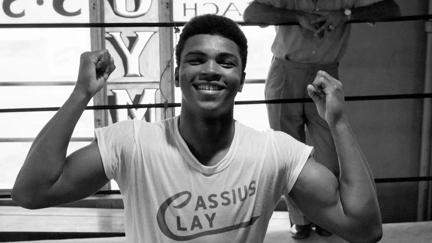 Young Cassius Clay smiling, raised hands, wearing Cassius Clay t-shirt