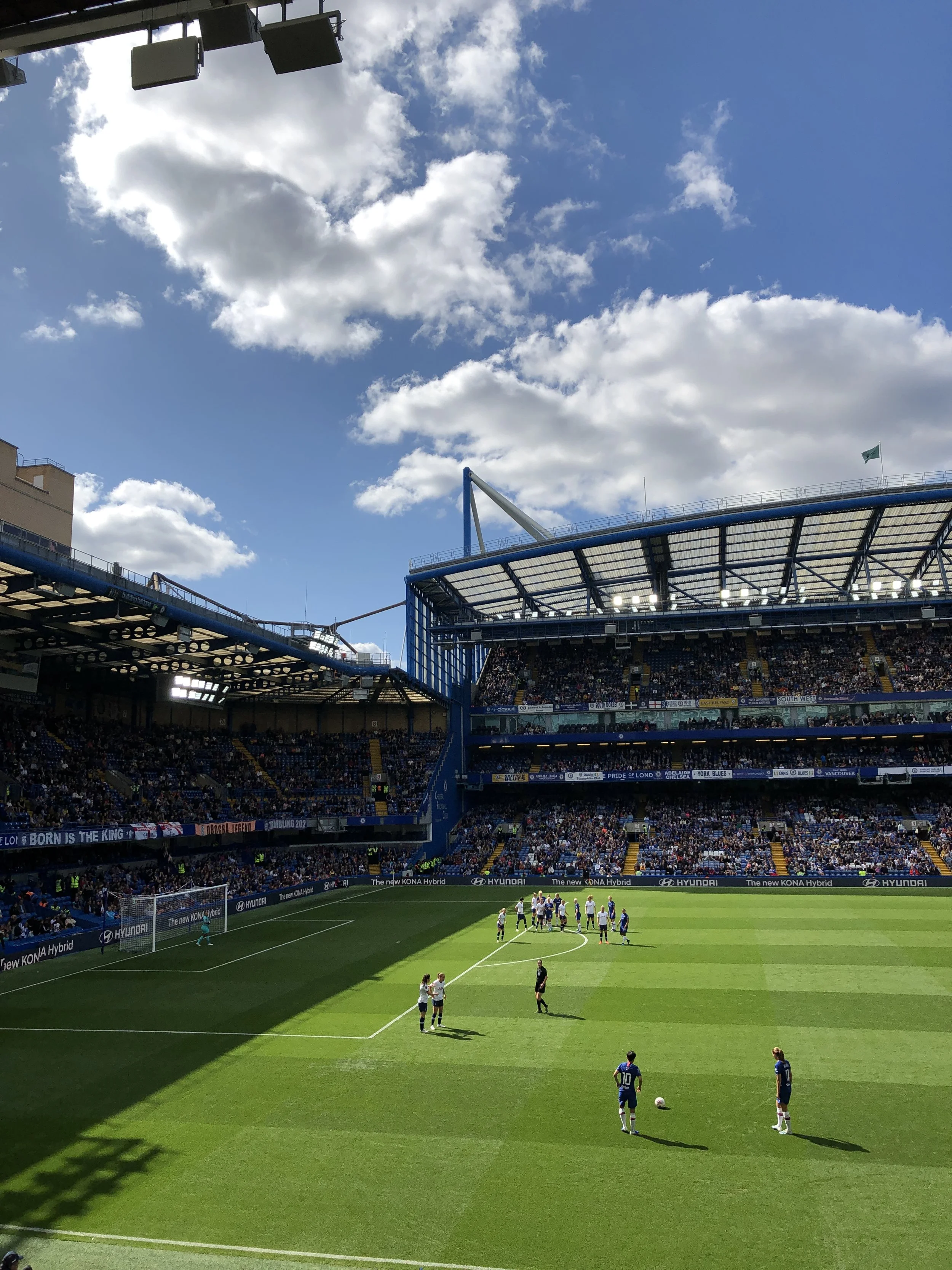 A soccer game in progress at a packed stadium on a sunny day with some clouds in the sky. Players are on the field near the penalty box, preparing for a free kick, with spectators watching from the stands.