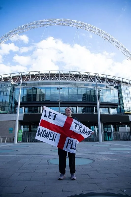 Person standing outside a modern building holding a sign that says "Let Women Play" with a red cross.