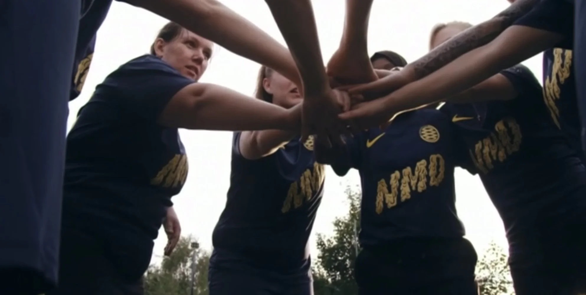 A group of women in dark sports uniforms forming a circle with their hands stacked together during an outdoor team huddle.