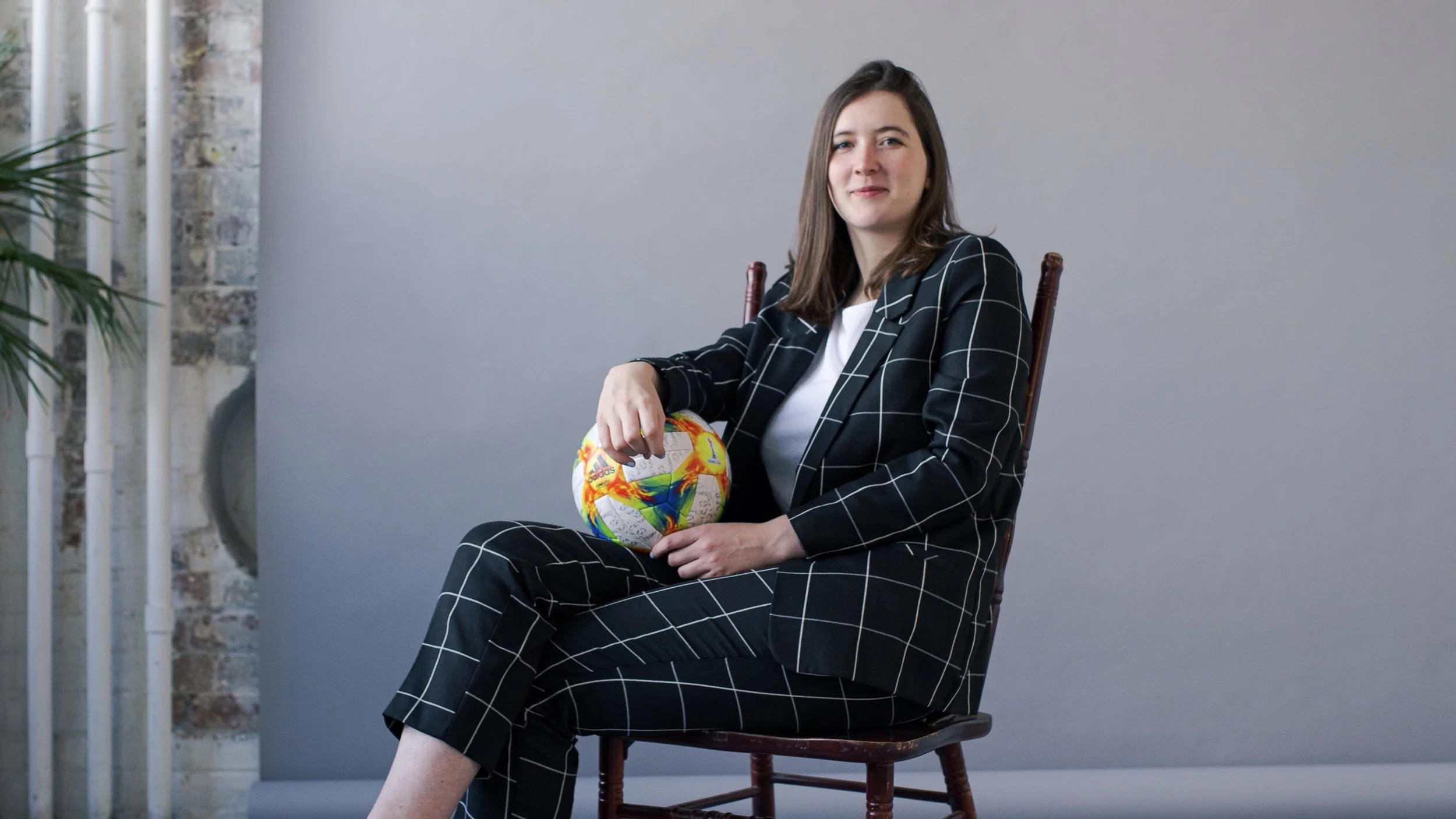 A woman in a black and white checkered suit sitting on a wooden chair, holding a colorful soccer ball, in a studio setting with a plain light gray background and part of a brick wall with a green plant on the side.