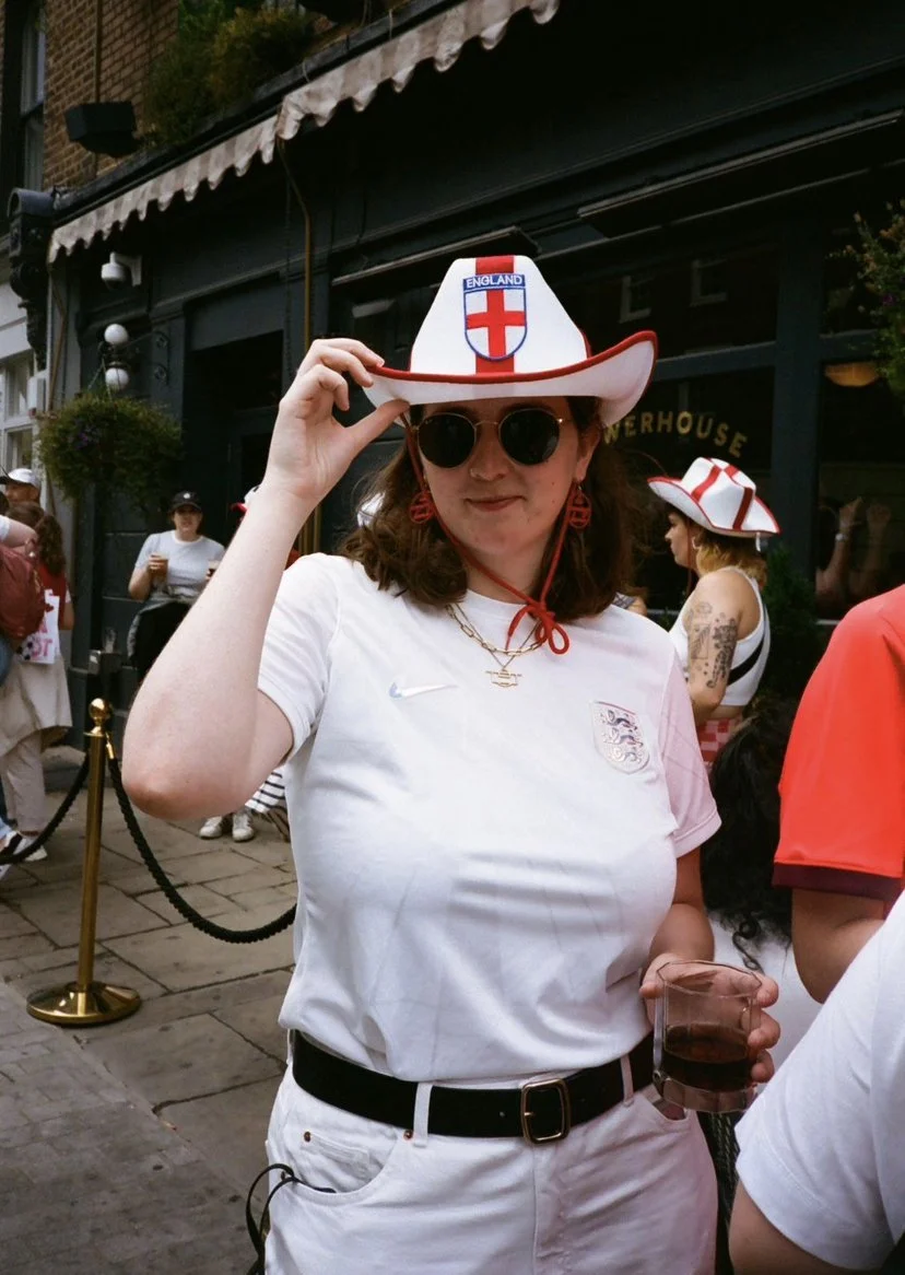A woman wearing a white England football jersey, round sunglasses, and a white cowboy hat with an England football crest, standing outside at a gathering.