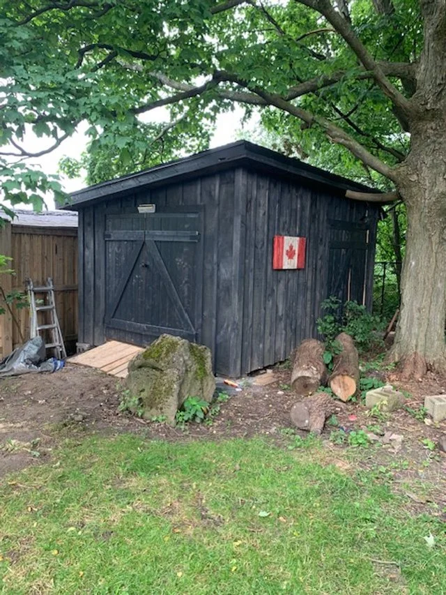 A small black wooden shed with a Canadian flag decoration on its side, surrounded by trees and logs, in a backyard with grass and a dirt area.