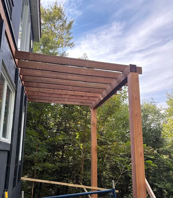 Wooden pergola with horizontal slats over a garden area next to a black house with siding, surrounded by green trees under a partly cloudy sky.