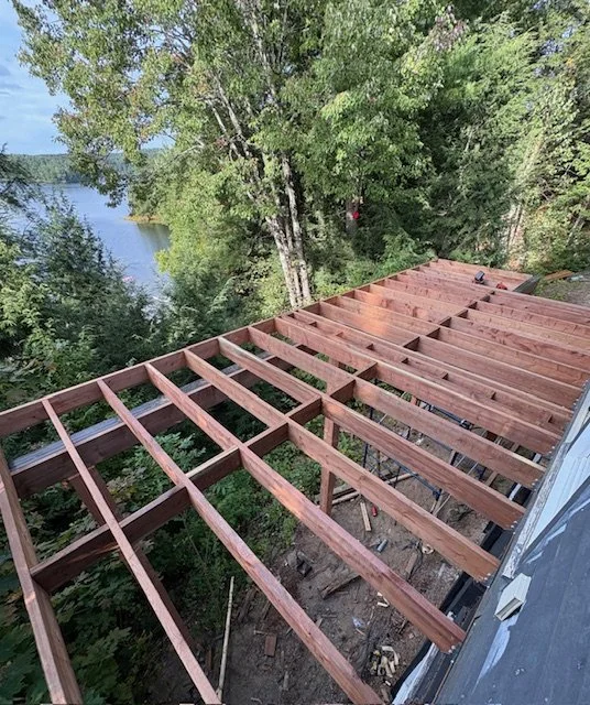 View of a wooden deck under construction with evenly spaced wooden beams, surrounded by lush green trees and overlooking a body of water.