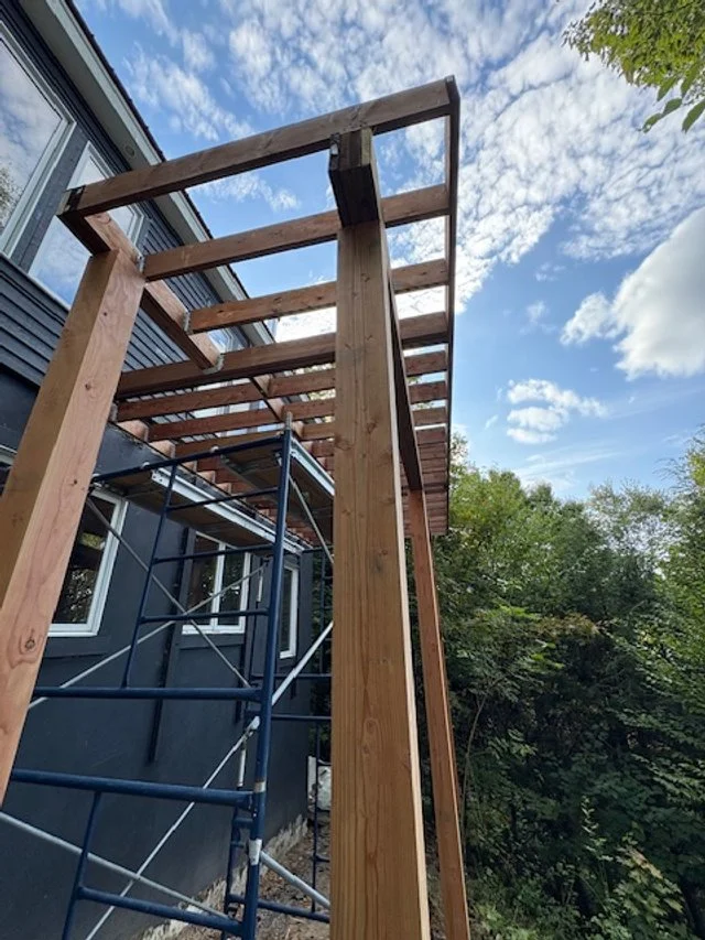 Wooden deck frame under construction outside a house with scaffolding, against a blue sky with scattered clouds and green trees.