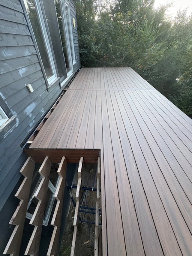 New wooden deck attached to the side of a house with blue siding and multiple windows, overlooking trees.