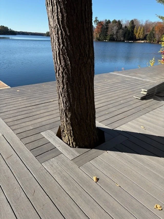 Tree growing through a wooden deck by a lake, with some stacked wood in the background during daytime.