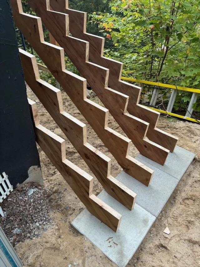 Wooden slab staircase under construction, with concrete foundations and a ladder in the background, outdoors surrounded by greenery.